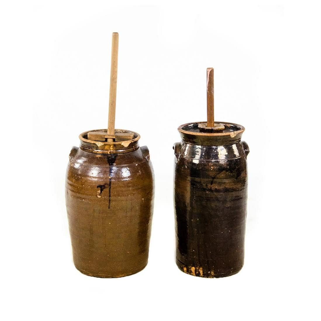 Pair of Antique Stoneware Butter Churns: A pair of antique stoneware butter churns. Each piece features a dark brown glaze and has an accompanying wooden pole. Condition: Wear consistent with age and history, including chips to each. Dimensi