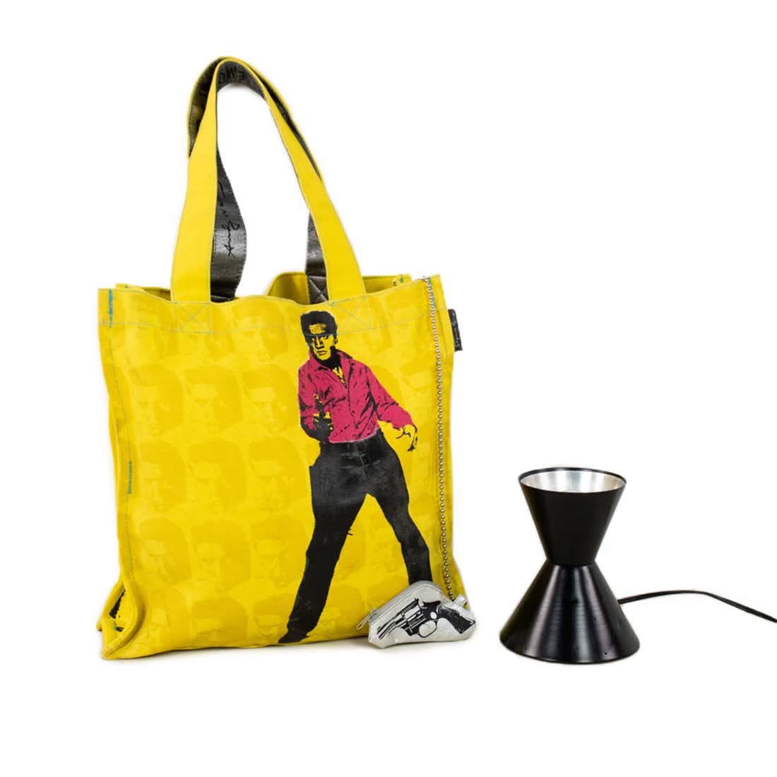 Andy Warhol Yellow Tote and Mid-Century Uplight Lamp: This lot includes an Andy Warhol yellow tote bag featuring a male figure in pink holding a gun with a small gun shaped keychain coin purse on the bag and a small metal uplight lamp. The uplight is unt