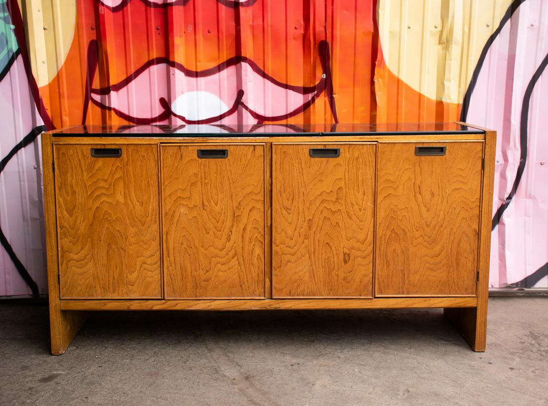 Mid-Century Modern Oak Veneer and Black Glass Credenza: Mid-Century Modern oak veneer panel and black painted glass top console credenza cabinet having four cabinet doors with three interior drawers and hidden compartment storage. Features a black painted 
