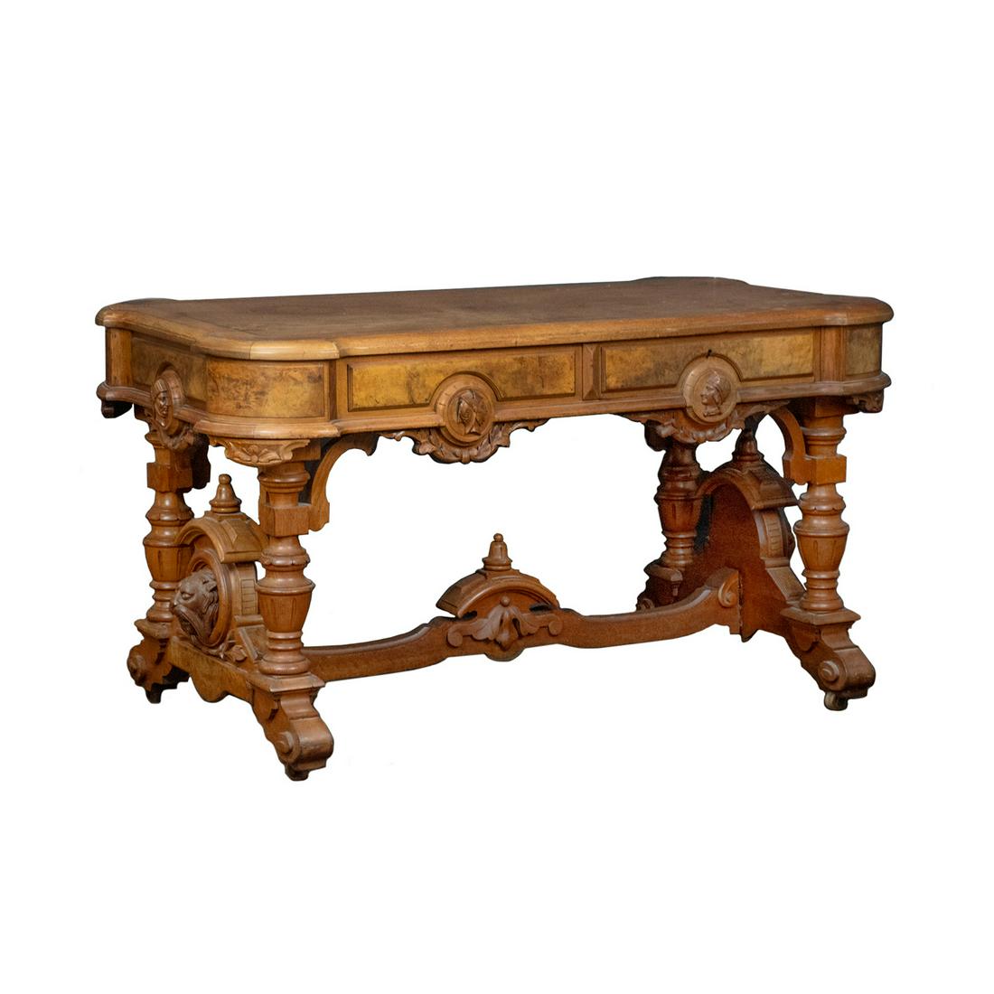 Antique Renaissance Revival Burl Walnut Library Desk: An antique Renaissance Revival burl walnut library desk. The table top features rounded corners, raised paneled sides, and a decorative apron with carved knight head profiles and foliate motifs. The b