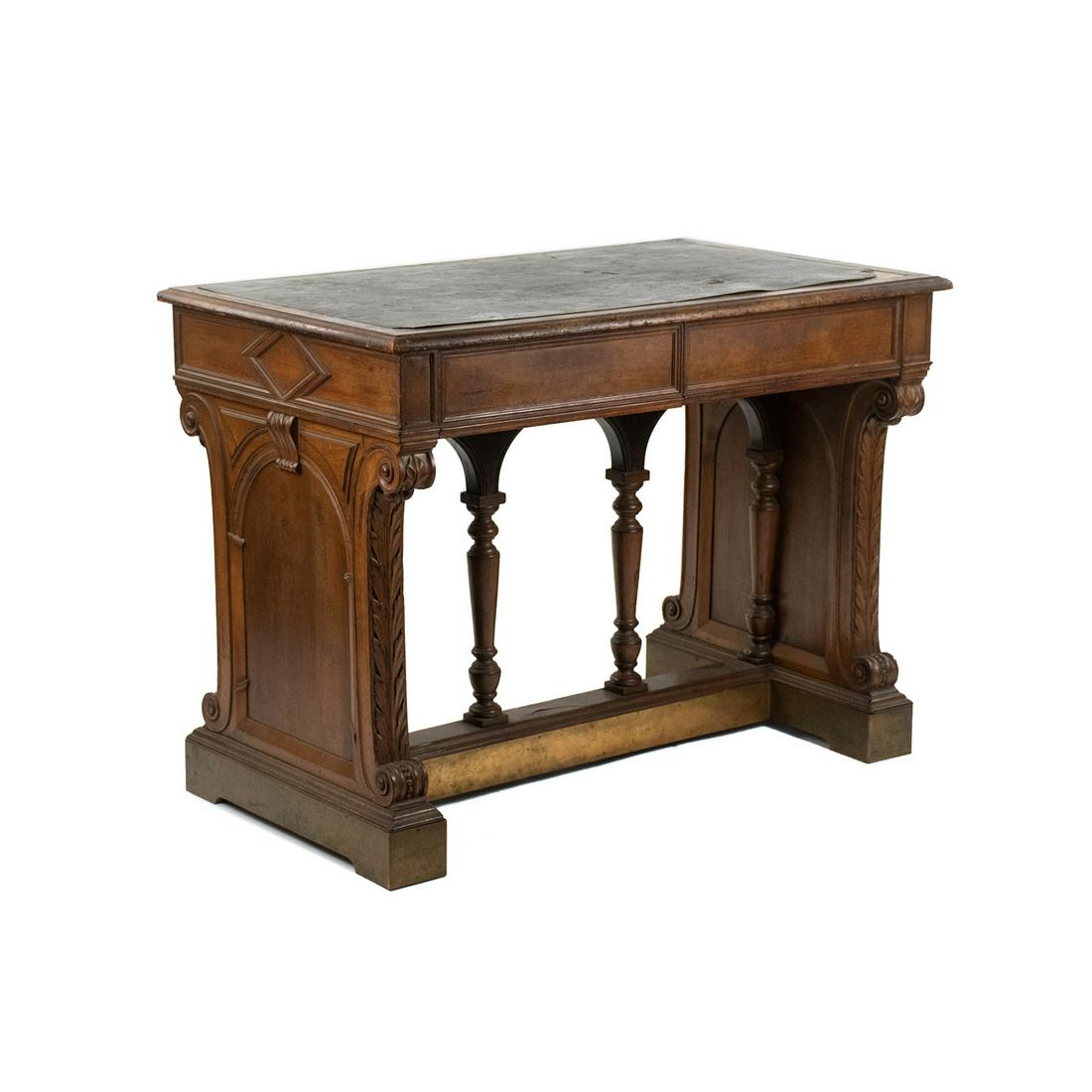 Italian Renaissance Style Writing Desk: An Italian Renaissance style writing desk having black leather top with two lower drawers flanked by scrolling acanthus carved columns resting atop a carved archway base with hammered brass border. Ma
