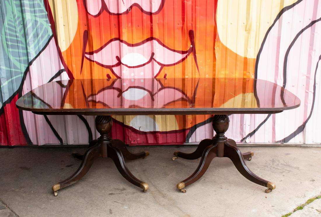 Sheraton Style Burl Oval Double Pedestal Dining Table: A large Sheraton style mahogany oval dining table with burl inlaid mahogany border and beveled edge resting atop a heavily carved double pedestal base having eight legs total with brass casters. Unmar