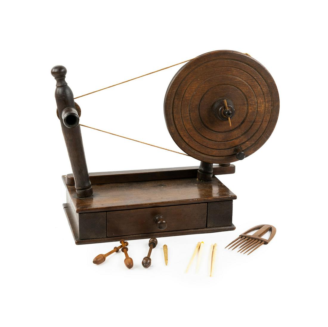 Spindle Type Lace Bobbin Winder: A bobbin winder on a stand having a turned grooved wheel with projecting handle connected by a belt to an upright spool holder, with a swing-out arm. Rests atop a rectangular base with a drawer contai