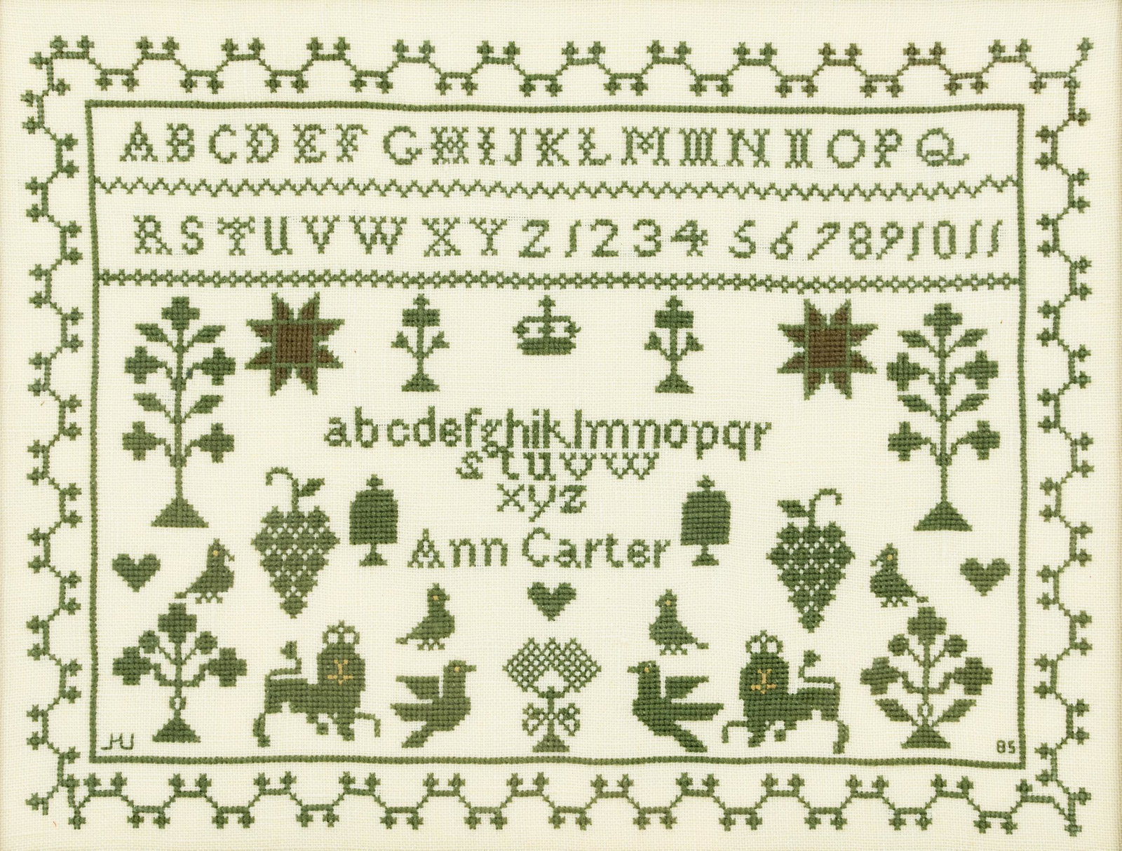 Ann Carter Traditional American Cross Stitch Sampler: An American cross stitch sampler with meander border, rows of letters and numbers, animals, foliage, and other symbols, and the maker's name stitched lower center. Stitched '85' lower right and illegi