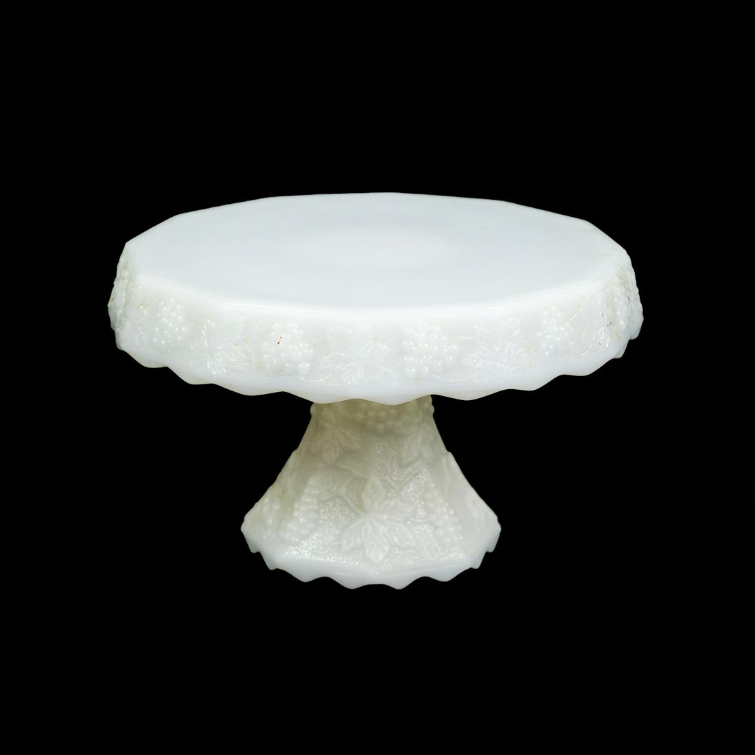Milk Glass Embossed Grapevine Cake Stand: A white milk glass round pedestal cake stand having an embossed grapevine motif around outer edge and on base.