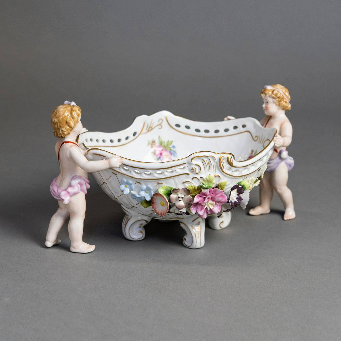 Meissen Porcelain Cherubs Basket Centerpiece: A Meissen porcelain centerpiece bowl having cherubs holding either side of a hand-painted floral basket with colorful floral appliques and gold accented design. Resting atop four decorated feet. Marke