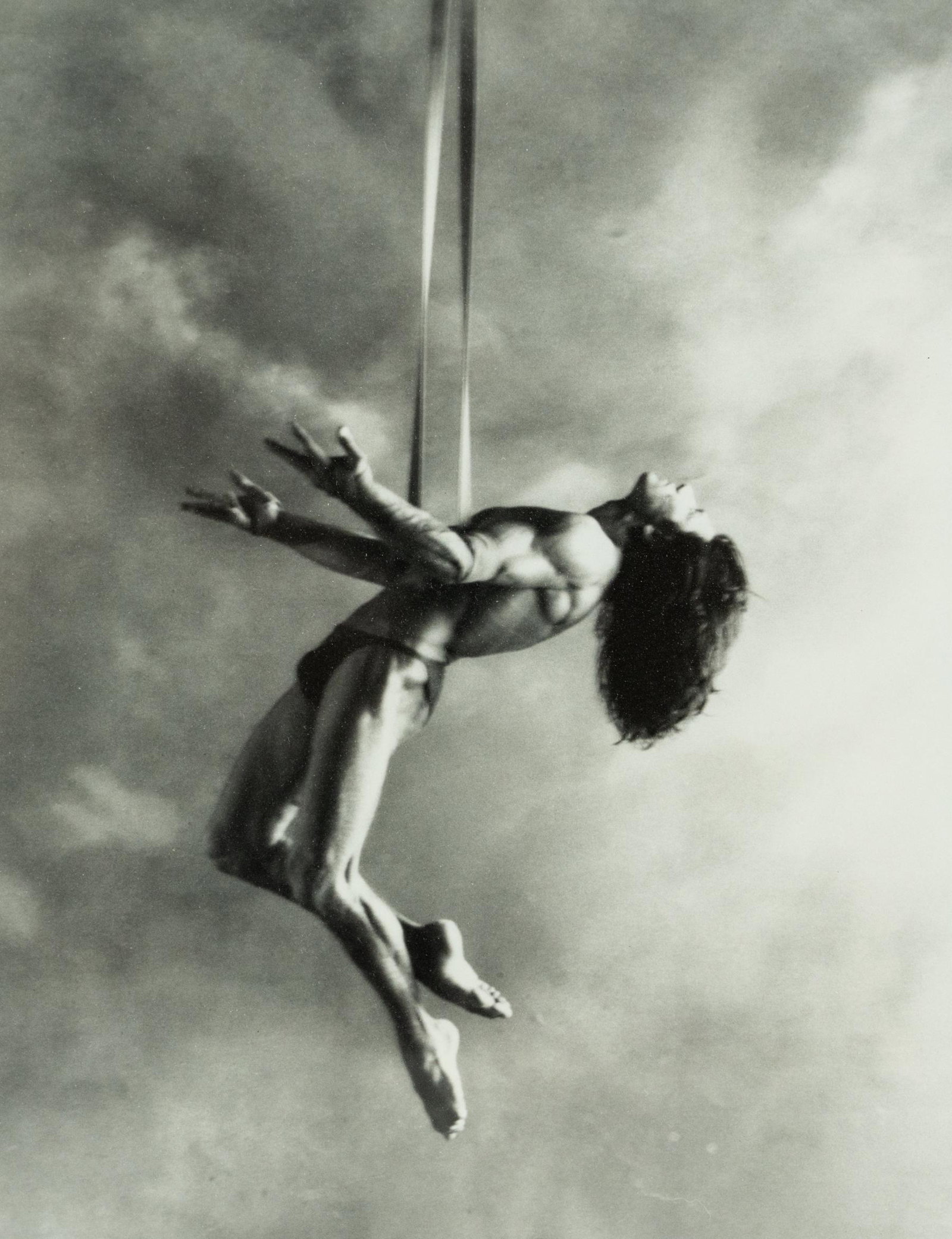 Aerial Silk Performer B&W Photograph: A black and white photograph of male aerial silk performer, suspended in an upward facing arched position with palms reaching up and legs gracefully positioned in the style of Herb Ritts (American 195
