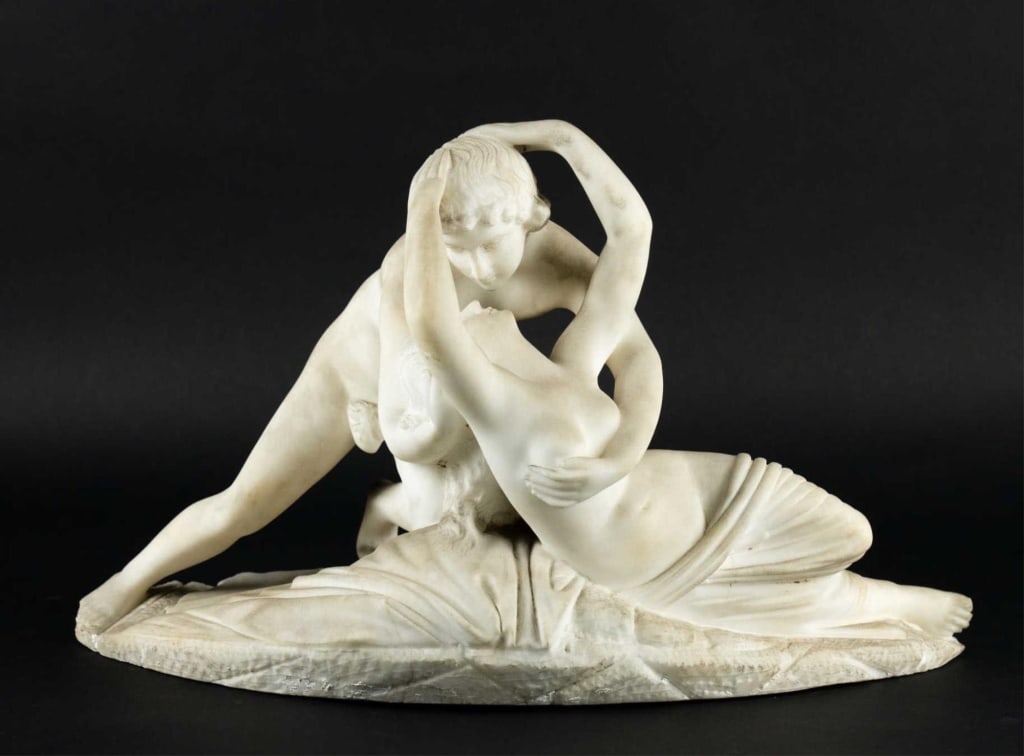 ITALIAN MARBLE CUPID AND PSYCHE AFTER CANOVA: Continental likely Italian late 19th–early 20th c. Carved white marble group of cupid and psyche after Antonio Canova; nude figures embracing on draped rocky base; integral oval plinth with toothed