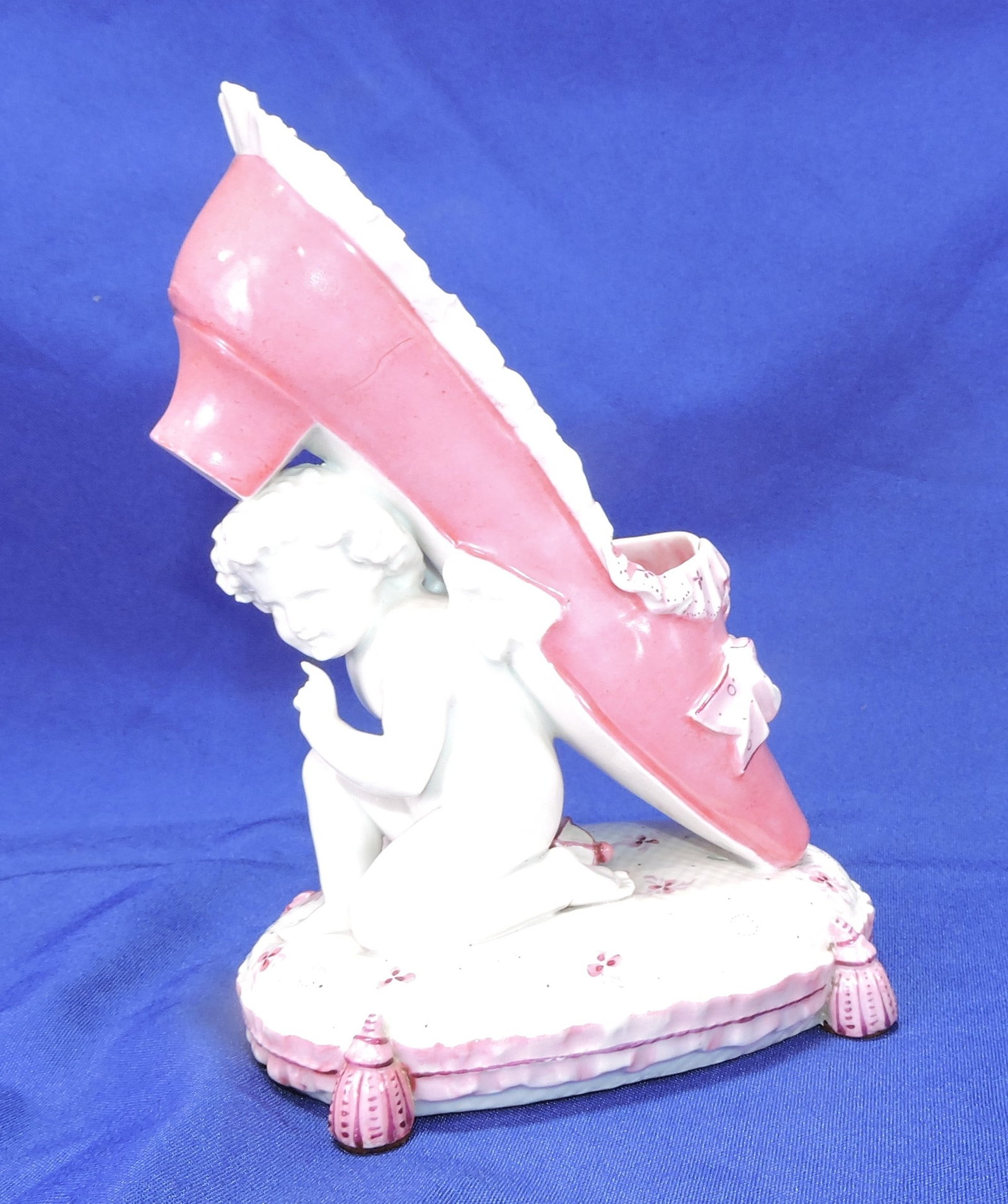 Porcelain figure of a winged cherub: supporting a pink shoe on an oval base, 8"