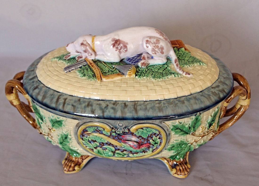 Minton majolica gun dog tureen and liner: modelled as an oval basket and cover, holly leaves to the sides, the holly branches forming handles at each end, an oval panel to each side, one depicting a running hare, the other a running pheasant,
