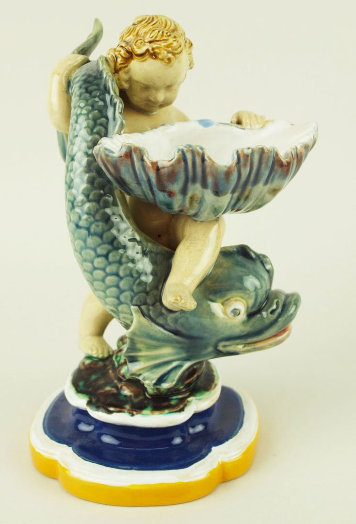 A Minton Majolica figural salt c.1858, modeled as a: A Minton Majolica figural salt c.1858, modeled as a putto straddling a grey-glazed dolphin and balancing a shell, glazed in mottled shades of grey and brown on a waisted quatrefoil pedestal glazed in