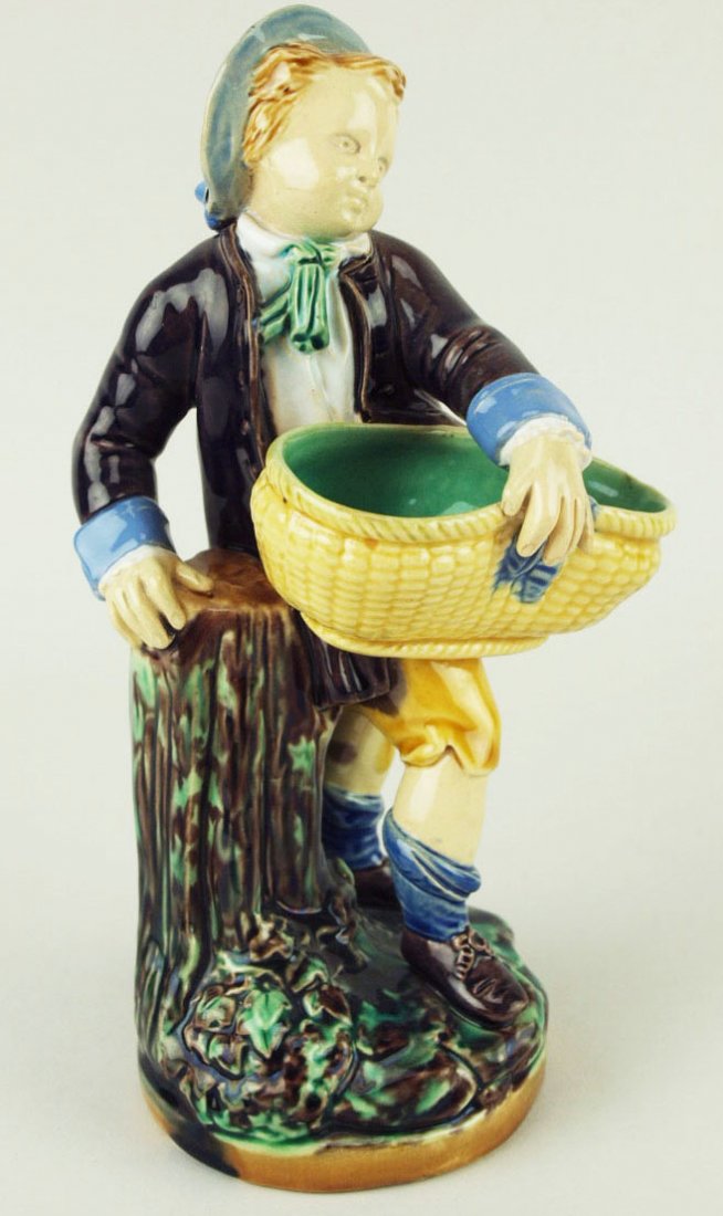 A Minton Majolica ‘Boy with Basket’ toothpick holder: A Minton Majolica ‘Boy with Basket’ toothpick holder c.1875, modeled as a young boy dressed in 18th century costume holding a yellow wicker basket and leaning against a mottled green and brown gre