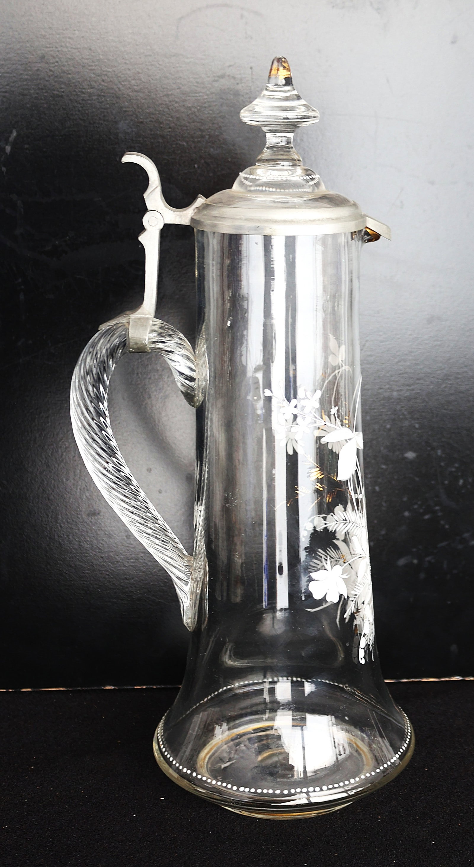 Vintage Claret Jug, a type of glass Wine Decanter, White Enamel Decoration, Twisted Glass Handle: Size: 15 1/2 in. height, 5 1/2 in. wide ; vintage claret jug, a type of glass wine decanter. It features a clear glass body with etched floral or leaf designs. The jug is topped with a metal hinged li