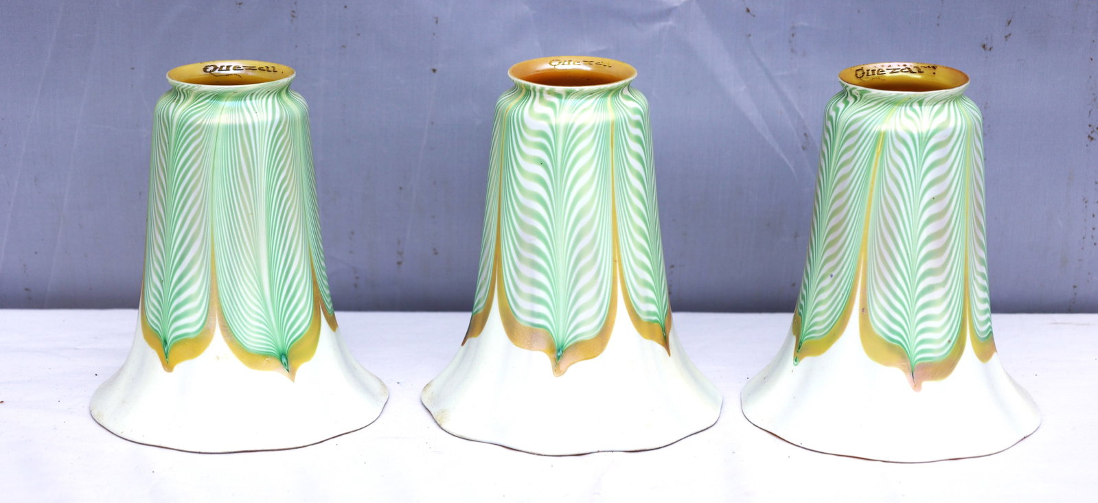set of three Quezal art glass lamp shades featuring a green pulled feather pattern tipped in gold: Size: 6 in. height, 5 1/4 in. diameter bell, 2 1/4 in fitter size ; set of three Quezal art glass lamp shades featuring a green pulled feather pattern tipped in gold