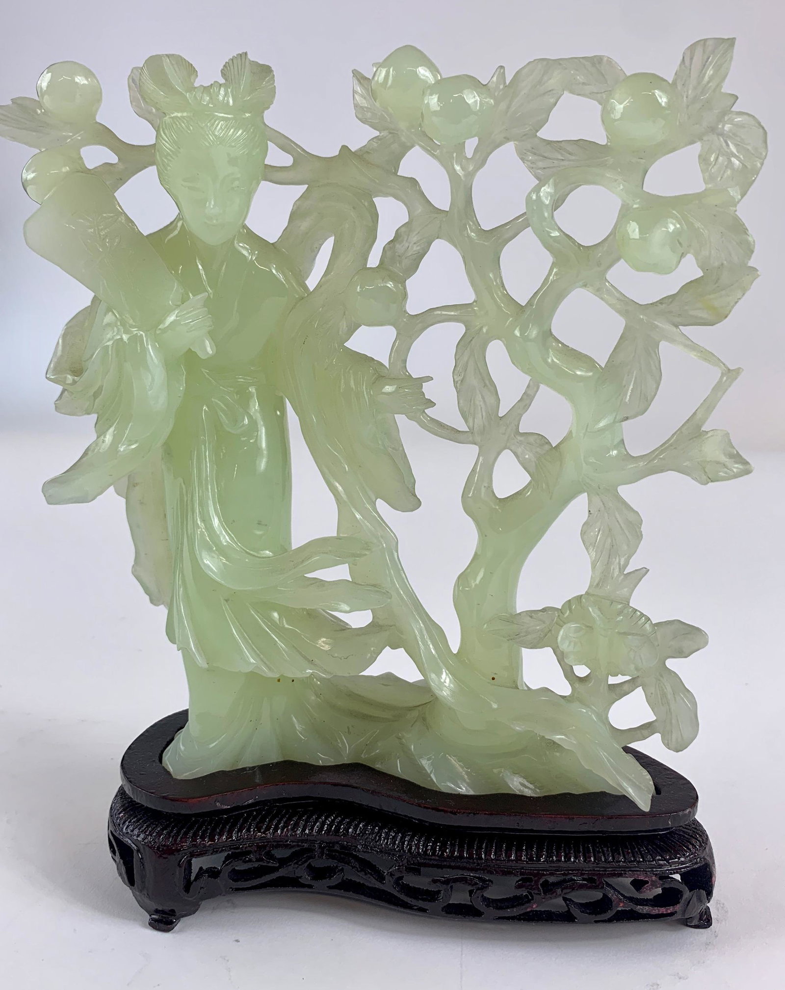 A Chinese Celadon Jade Guanyin Figural Carving on Stand: 20th century. A Chinese carving in celadon toned jade, depicting a Guanyin figure with fan, aside a fruit and branch pierced carved setting, atop carved rosewood stand; total ht. 9".