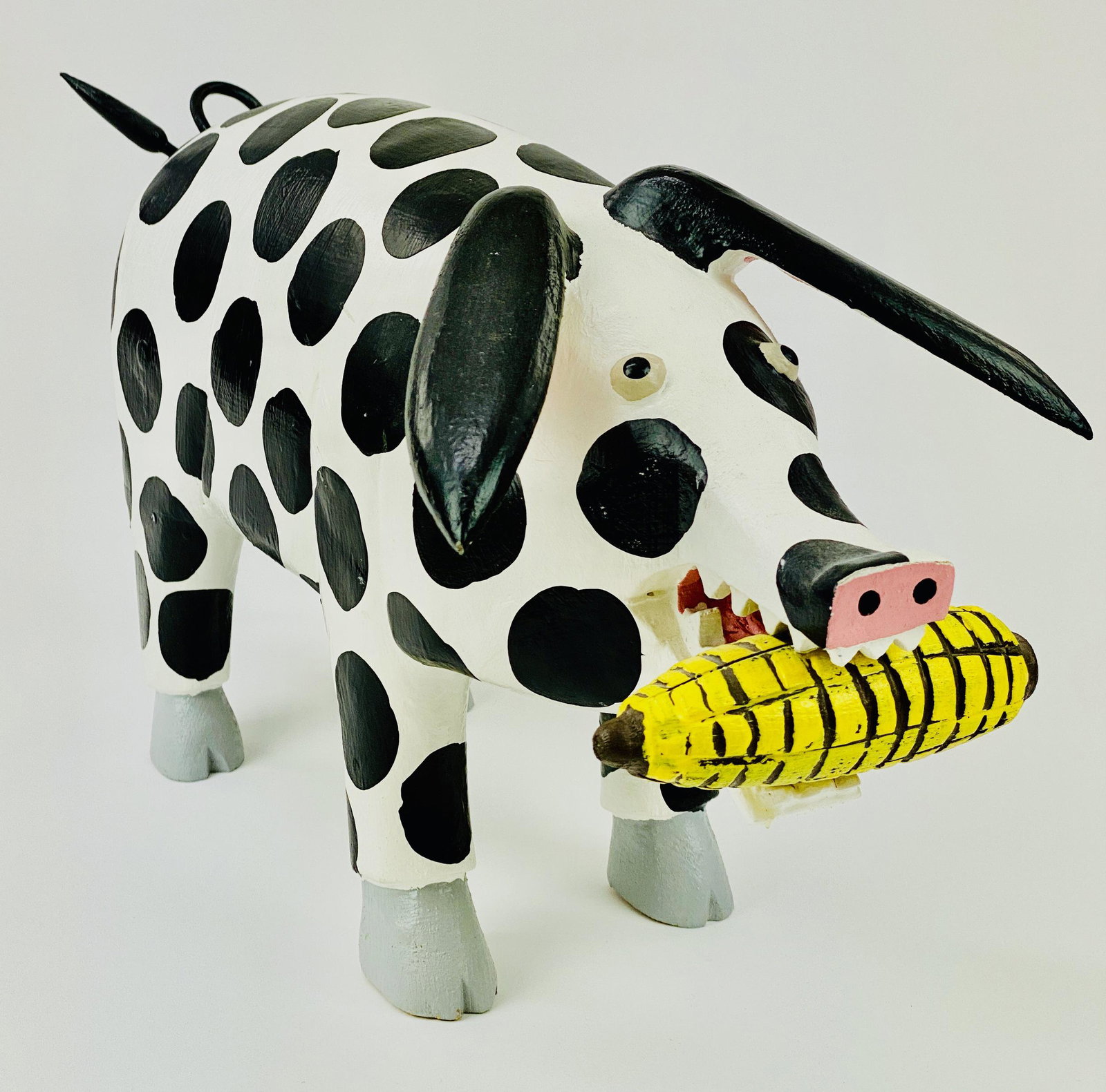 Folk Art Carved Pig by Lonnie and Twyla Money: A well executed carving by Lonnie and Twyla Money folk art featuring a pig holding a corncob in its mouth and a curly tail, signed in pencil on the underside of the hooves; 9.5" ht. x 16" wd. From th