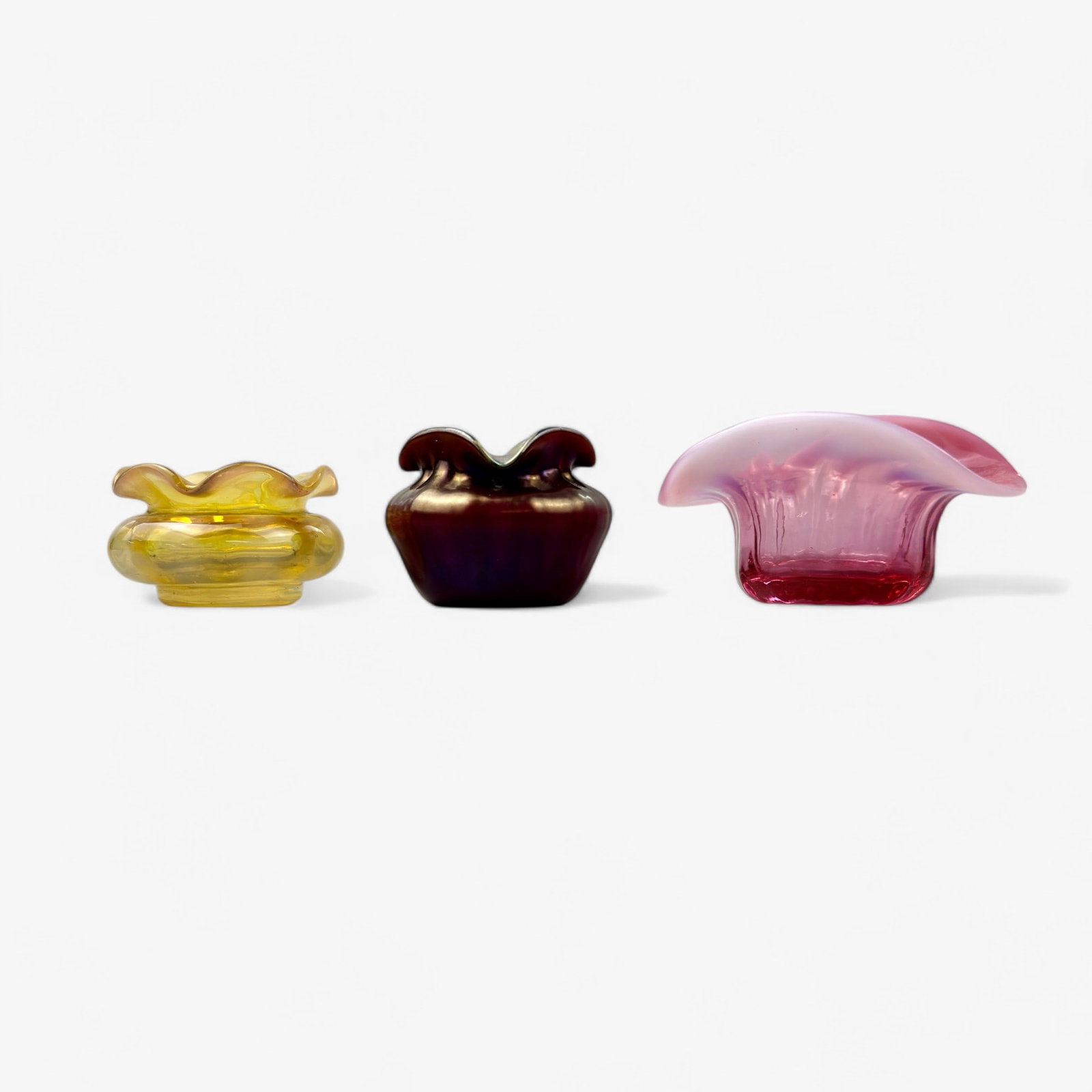 Group of Three Miniature Art Glass Bowls: One iriedescent vessel with flared rim by Douglas Nash, one yellow glass vessel with flared rim, unsigned, and one rose/pink art glass dish with flared rim. Height of Douglas piece: 1.5", height of ye