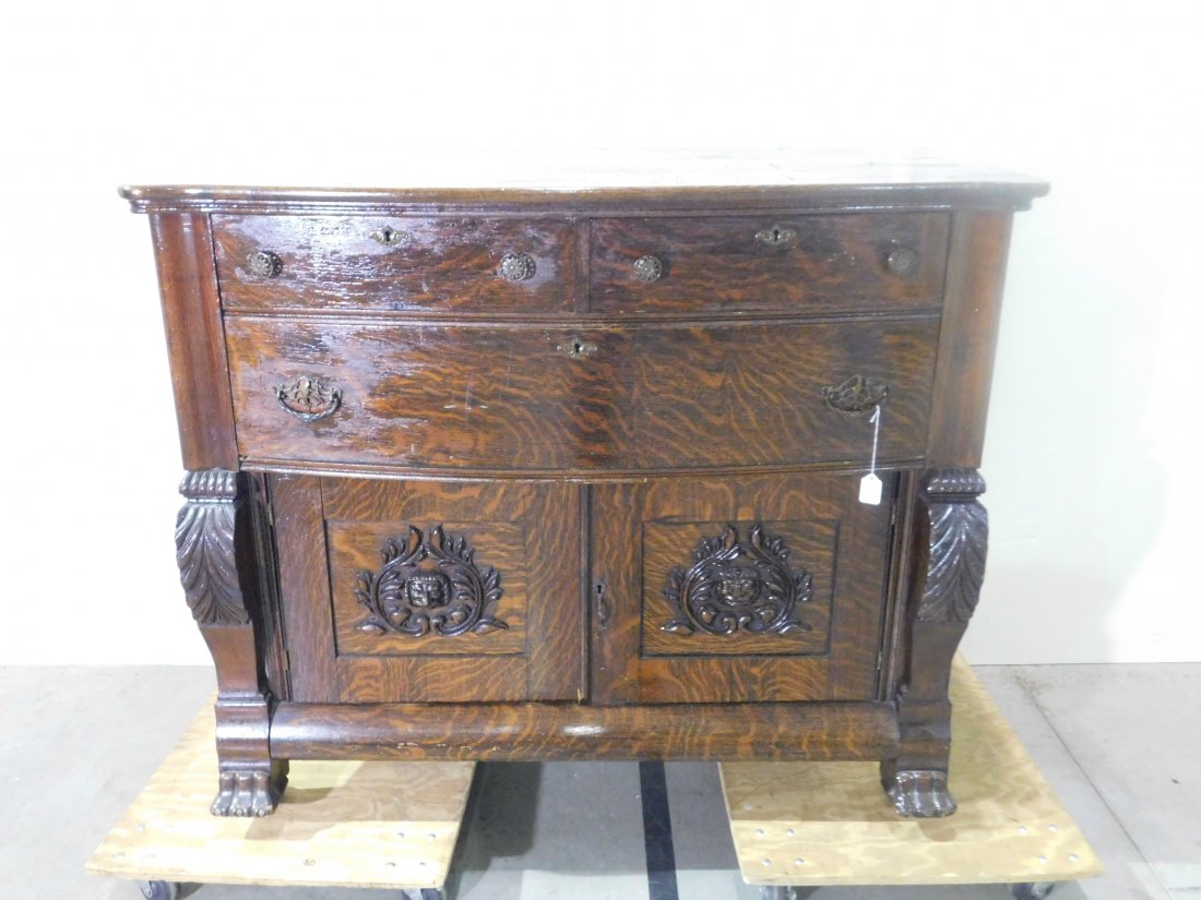 Ornate Oak Sideboard: Ornate oak bowfront sideboard with one lionshead and one man\'s head. 3 drawers, 2 doors, on wooden wheels. Approximately 48 inches wide, 40 1/2 inches tall, 23 inches deep. Overall good condition.