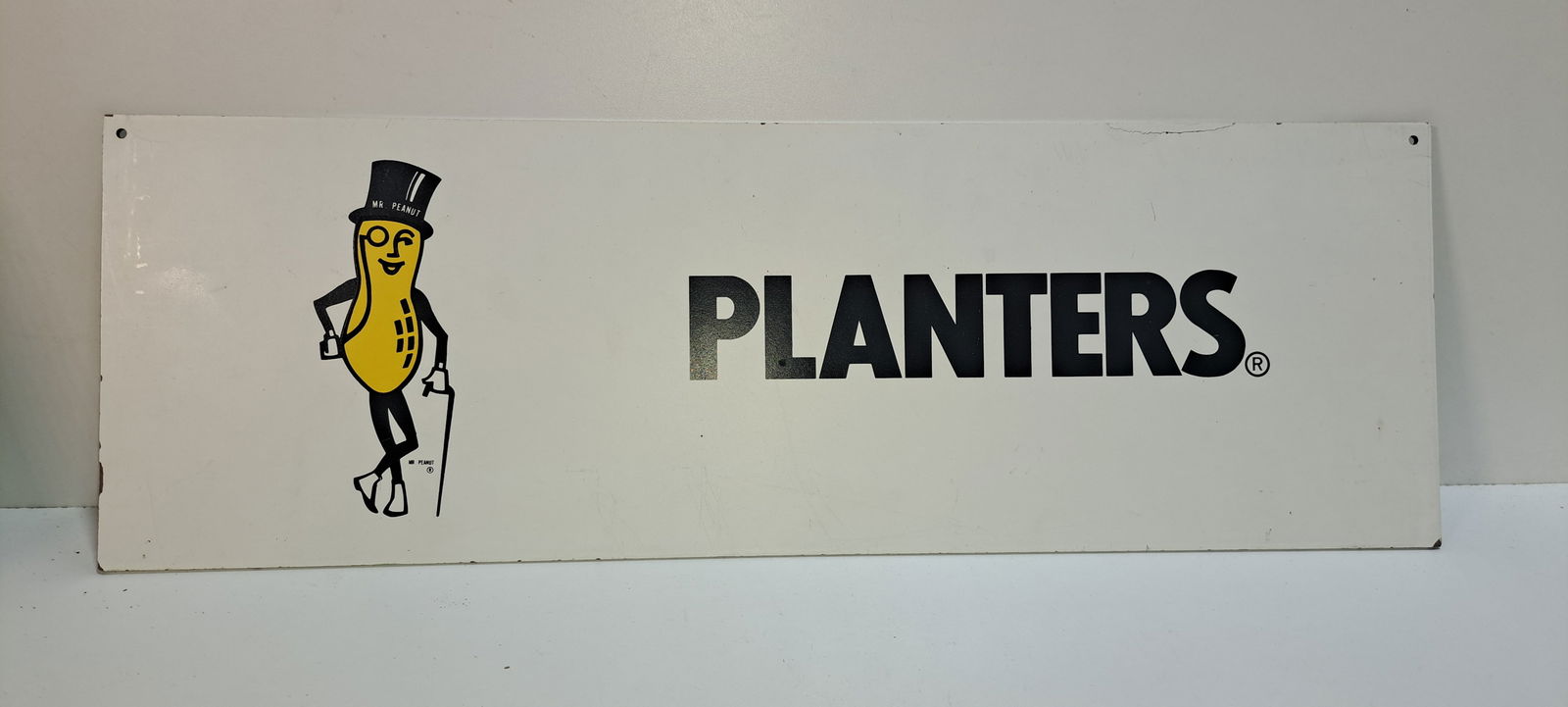 Planters/Mr. Peanut sign: single sided, masonite sign With Mr. Peanut, shows edge wear and small crack on top, approx. 8" x 24", Please note: if picking up, location is different than auction location. Please contact us
