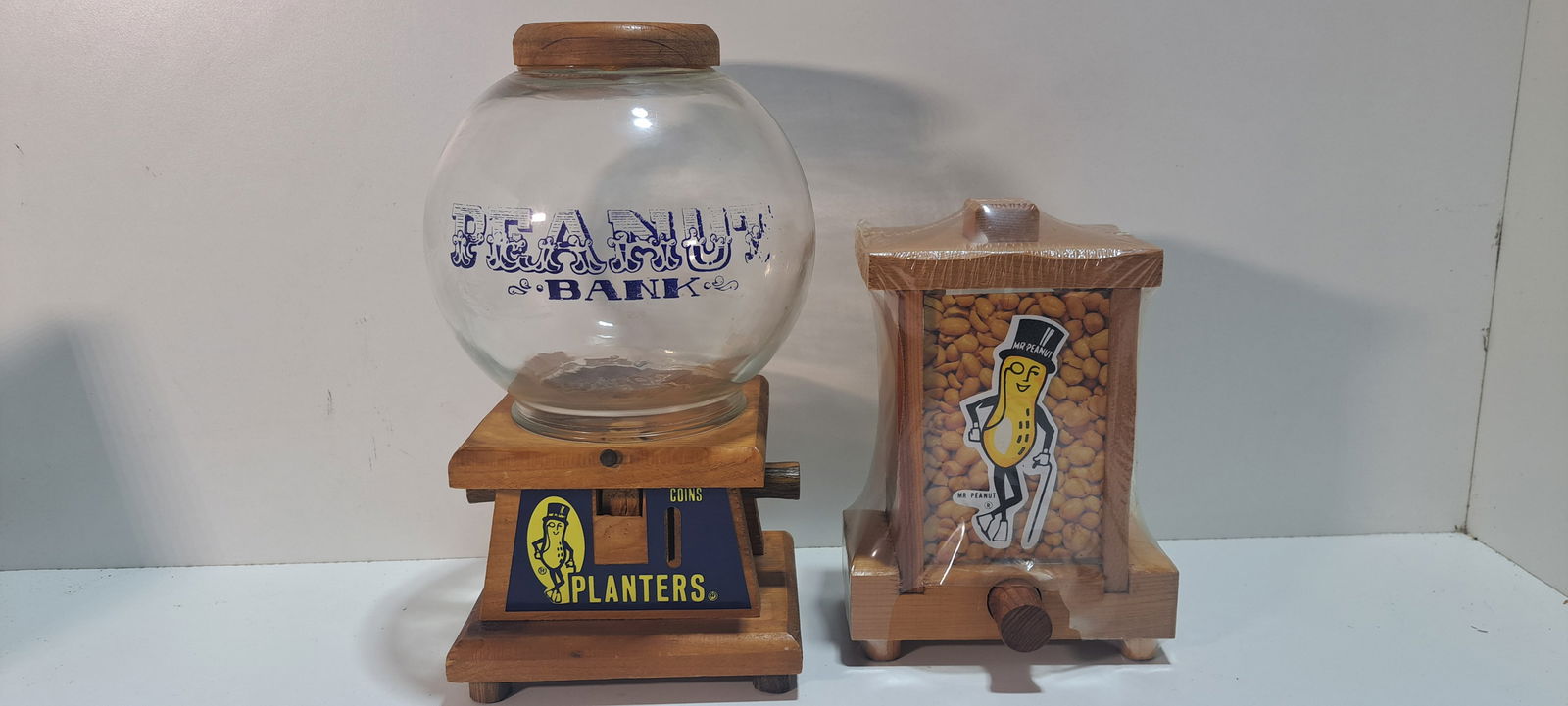 2 Planters Peanuts nut dispensers: 2 wooden and glass, Planters Peanuts, nut dispensers, larger one has wear to paint on glass, smaller one still in shrink wrap, unused, approx. 12.75" tall, and 9.25" tall, Please note: if picking up,