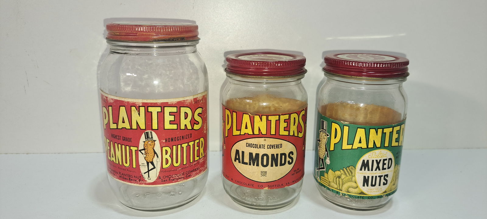 (3) Planters Peanuts glass jars: 3 small glass jars, metal screw on lids, paper labels, with Mr. Peanut, peanut butter, chocolate covered almonds, mixed nuts, minor wear, one at 5.25" tall, others 4.25" tall, Please note: if picking