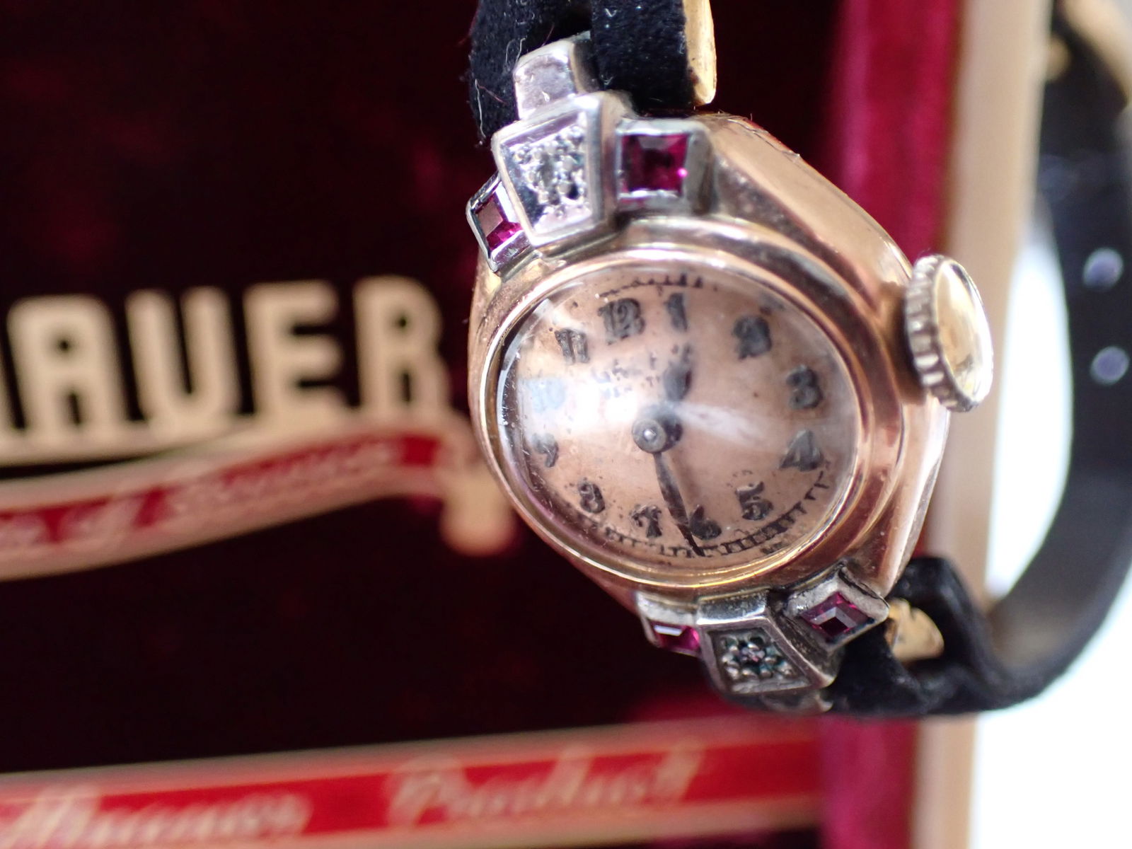 Woman's Gold Filled Wristwatch Face is marked Geneva: Case is marked The Gruen Watch Co. In a Wittnauer plastic case. The watch has ruby colored glass accents with leather strap. Writing on watch face is worn. Plastic case has some wear with a crack. The