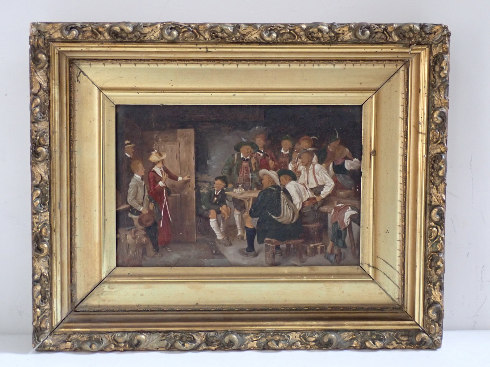 Bavarian Bar Scene Oil on Plank Painting: Painting depicts family opening door where a group of men are drinking at a table. Gold Gild Frame. Marked on back Q plank is F.V., Deeringer, Mittastrast & the number 4960. Frame by Charles W. Dennis