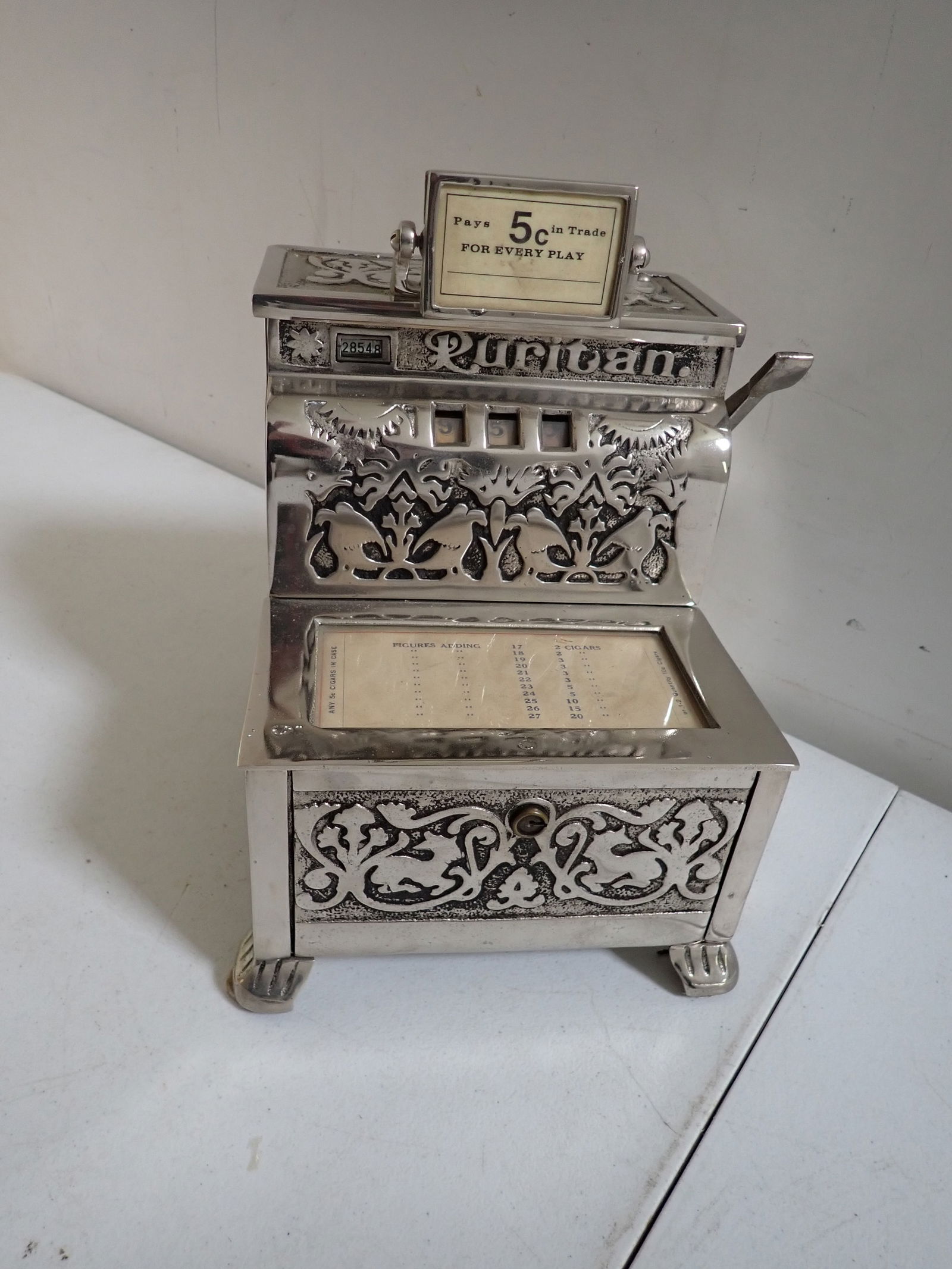 Caille Puritan 5¢ Trade Stimulator: Nickel Plated Silver Trade Stimulator. Pivoting Label says 5¢ in Trade For Every Play. Numbered 28548. Has windows for 3 numbers. The numbers move when nickel is deposited and lever is pulled but don