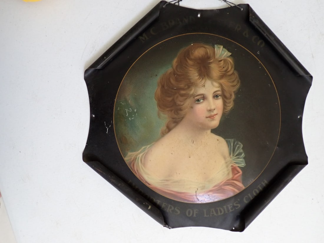 M. C. Brandstetler and Co. Chicago Importers of Ladies Cloth metal sign: Circular design with Image of Victorian woman and name below her is Lenore. Signed B. Geyer. Four metal edges are curled over. Measures 14 1/2" x 14 1/2". Has some scuffing and wear and cracking but i