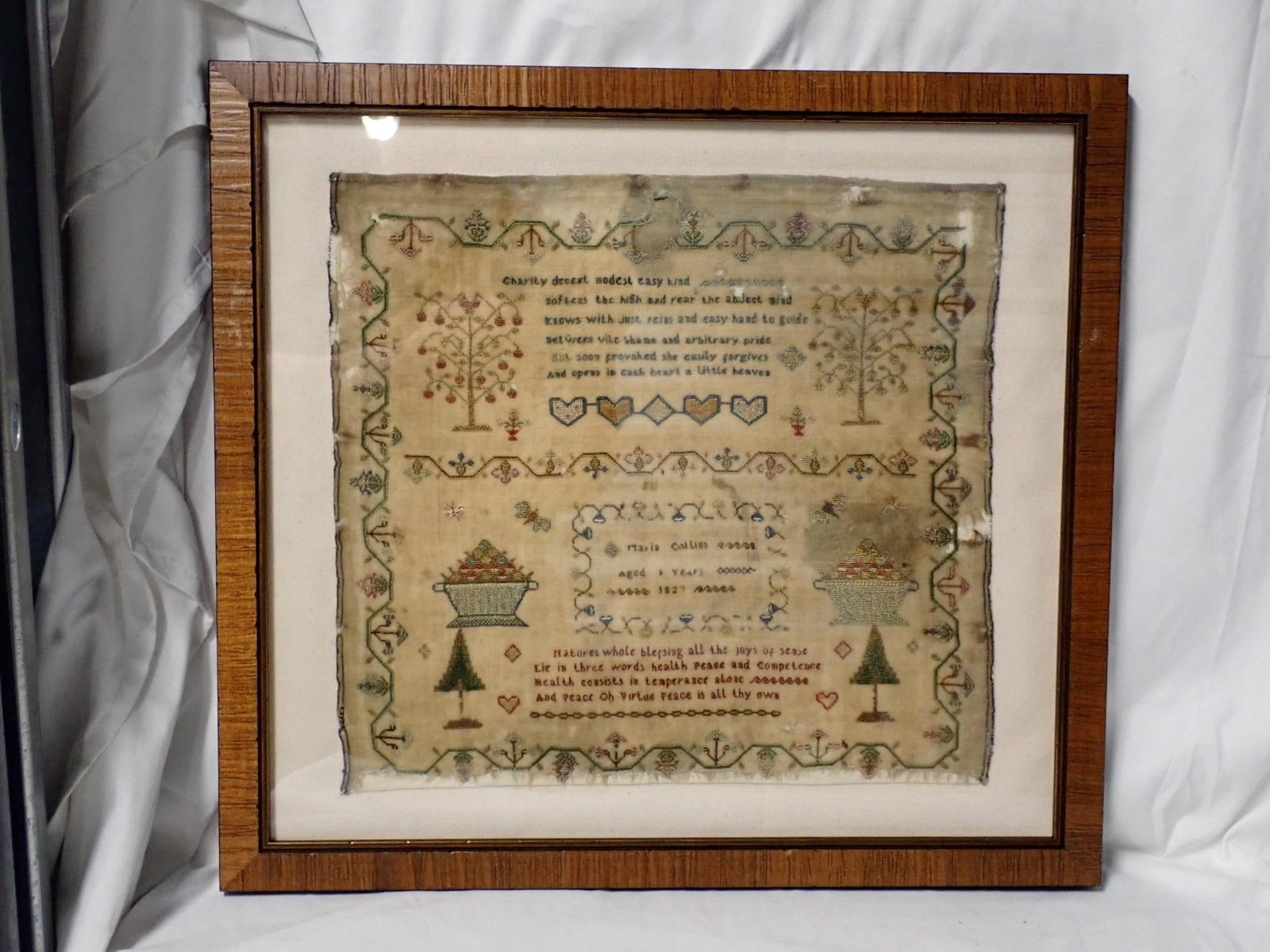 1822 Maria Collins Needlework: Maria Collins Aged 9 (?) Years 1822 (?) Verse on top and bottom motif is hearts trees baskets. Border geometrical vines and flowers. Fabric loss, staining and discoloration Frame 16" x 17 1/2"