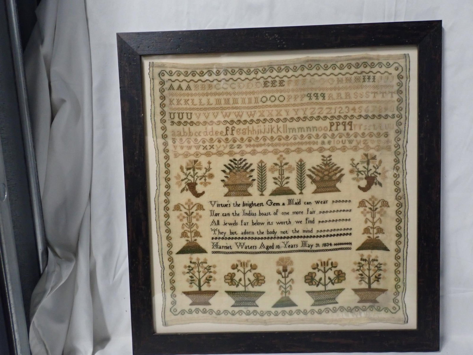 1834 Harriet Waters Sampler: Harriet Waters Aged 10 Years May 31, 1834 Alphabet and numbers. Verse in center Motif Flowers in baskets. Vining and circles border. Some fabric discoloration. Some fabric loss. Frame 17 1/4" x 16