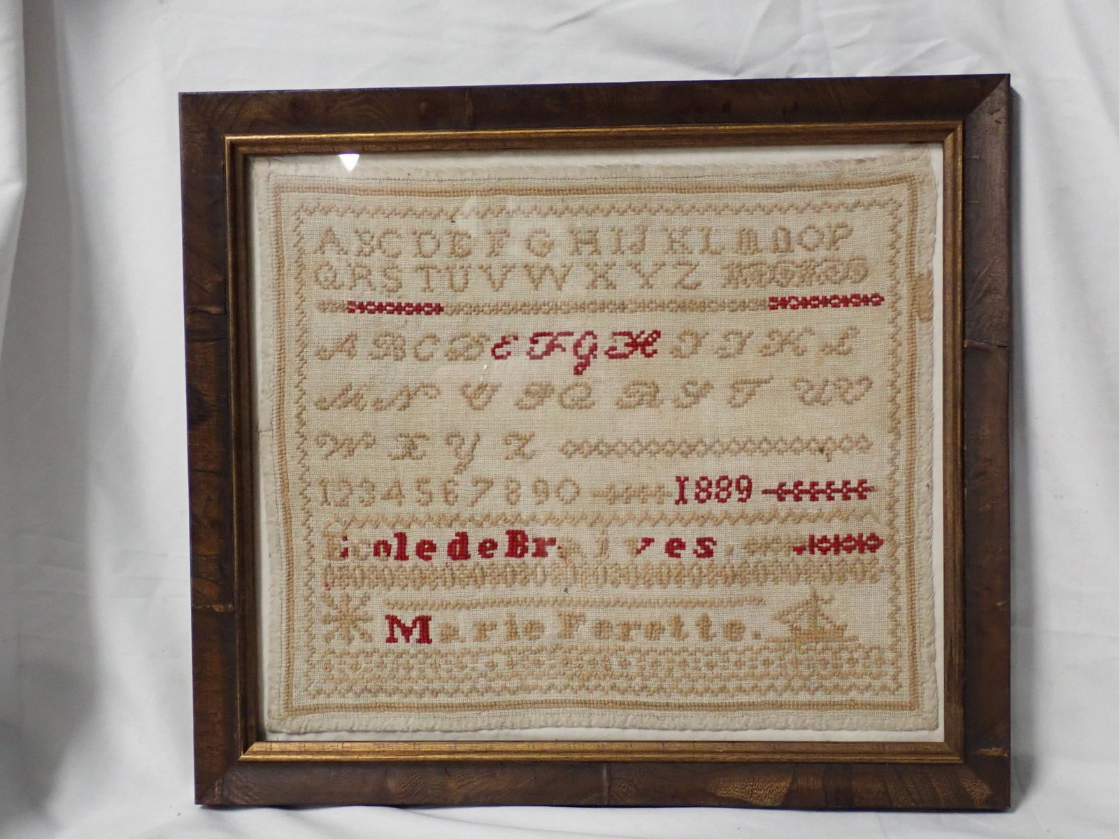 1889 Marie Ferette Sampler: 1889 Ecole Braives Marie Ferette. Alphabet and numbers. Small ship after name. Geometrical border. Slight stain on edge. Frame 13 1/2 x 15 1/2. Sampler 10 3/4 x 12 3/4