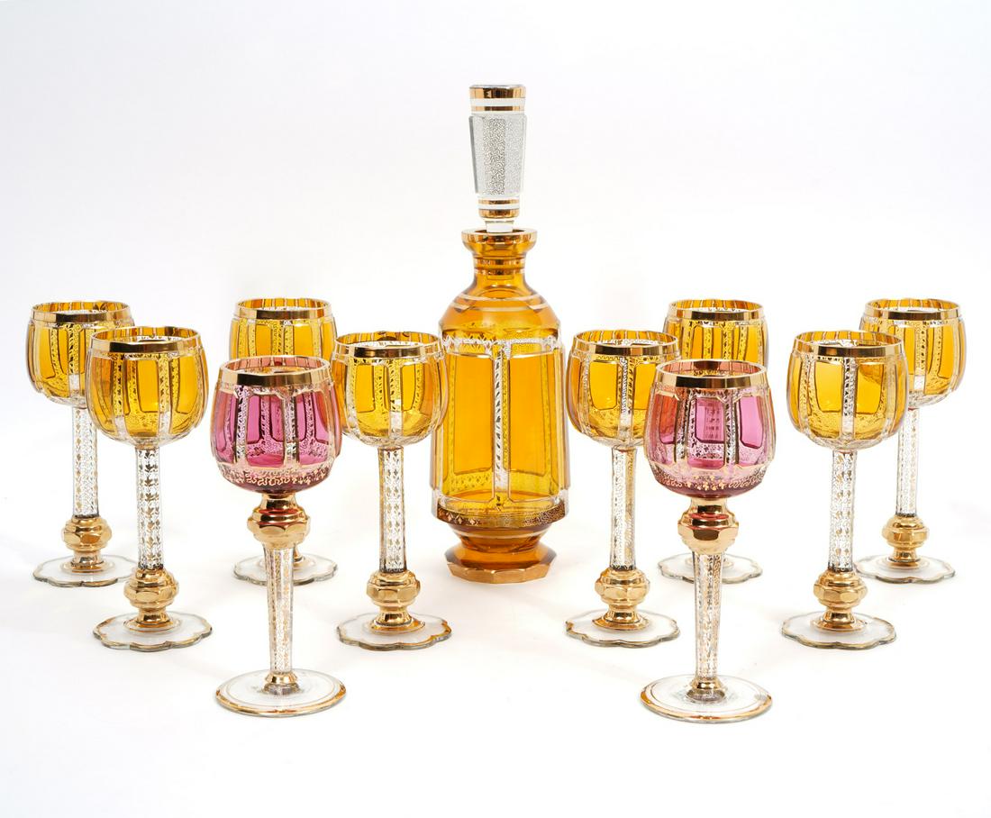 Moser-Style Gilt Decanter and 10 Wine Goblets: Moser-style gilt leaf pattern decanter with 10 wine goblets. Two goblets are cranberry red and 8 are gilt leaf pattern. Decanter measures 14.25"H x 4"W. Goblets measure 8"H x 3"W. Condition report: Ch
