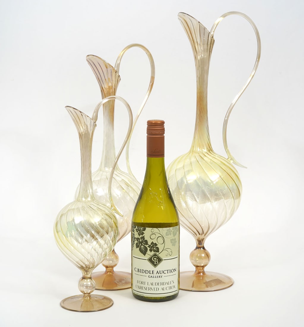 Three Murano Italian Art Glass Swirl Pitchers: Three Murano Italian art glass pitchers in champagne colored swirled glass with long slender necks, applied curled handles and circular base. Measures 13 3/8"H x 4.75"W; 16.75"H x 5.75"W and 19"H x 6"
