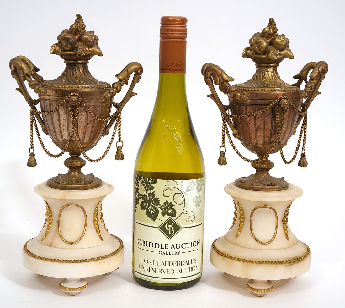 Pair of Bronze and Cream Marble Garnitures: Pair of marble and bronze garnitures have double handled urns, fruit-style finials and draped chains. They are mounted on cream-colored marble pedestals with tripod footed base and bronze beaded accen