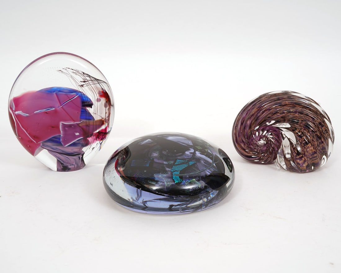 Three Assorted Art Glass Paperweights: Three assorted art glass multicolored paperweights includes an oval shaped piece with flat bottom (5"H x 4"L x 1.25"D); circular piece (5"L x 2"D); swirled sea shell shape (3.25"H x 2 3/8"W x 4"L). Th