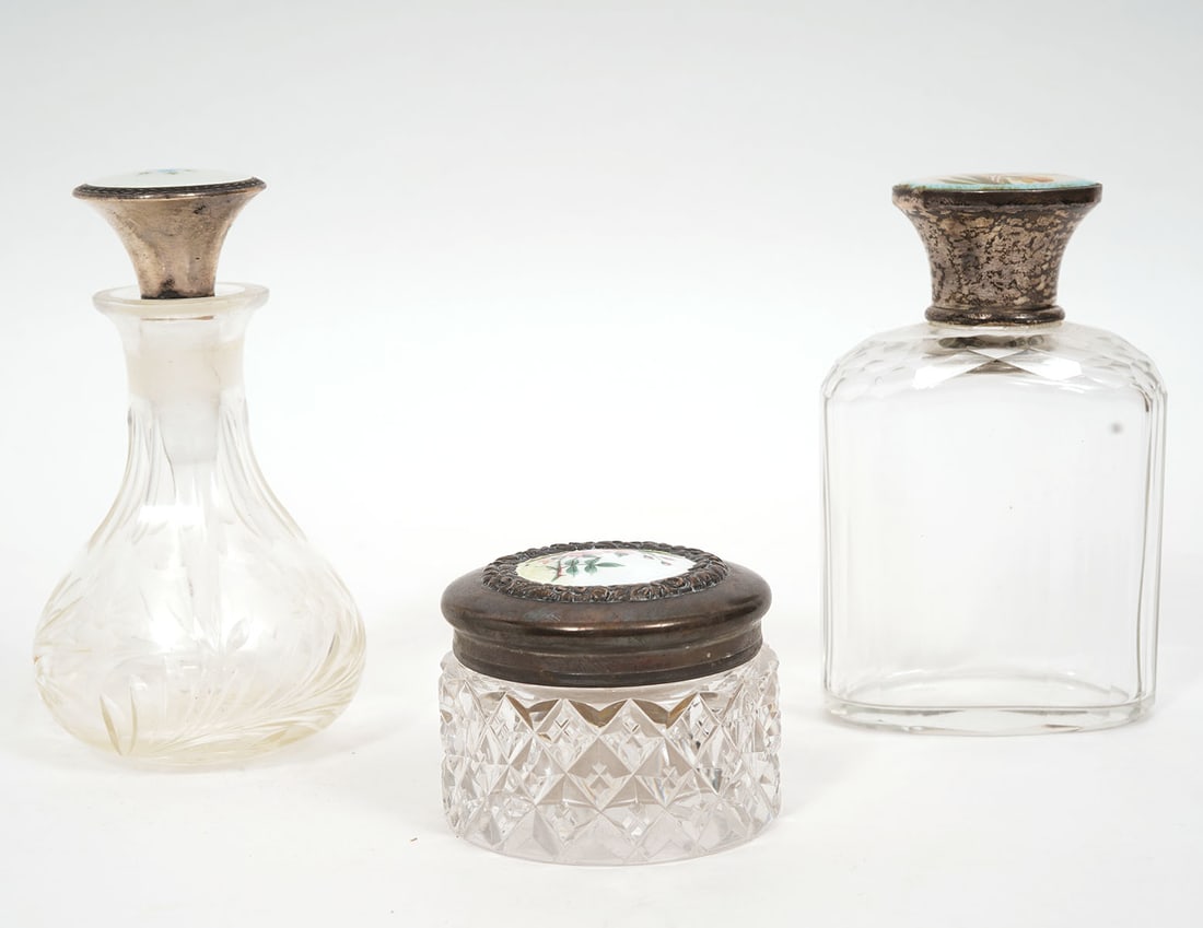 3 Cut Glass Bottles with Enameled Silver Lids: One American cut glass jar with silver lid decorated with enameled flowers measures 1.5"H x 2"W. One English etched glass perfume bottle has blue enameled flowers on lid and measures 4.75"H. One oval