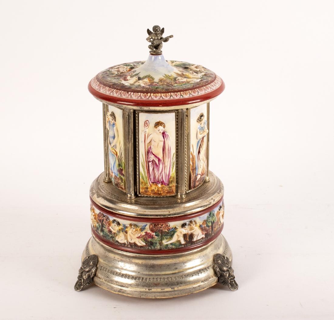 A Capo di Monte style musical cigar dispenser, with: A Capo di Monte style musical cigar dispenser, with cherub finial, the button operated clockwork mechanism opening to reveal six cigar apertures, on satyr mask feet, 27cm high