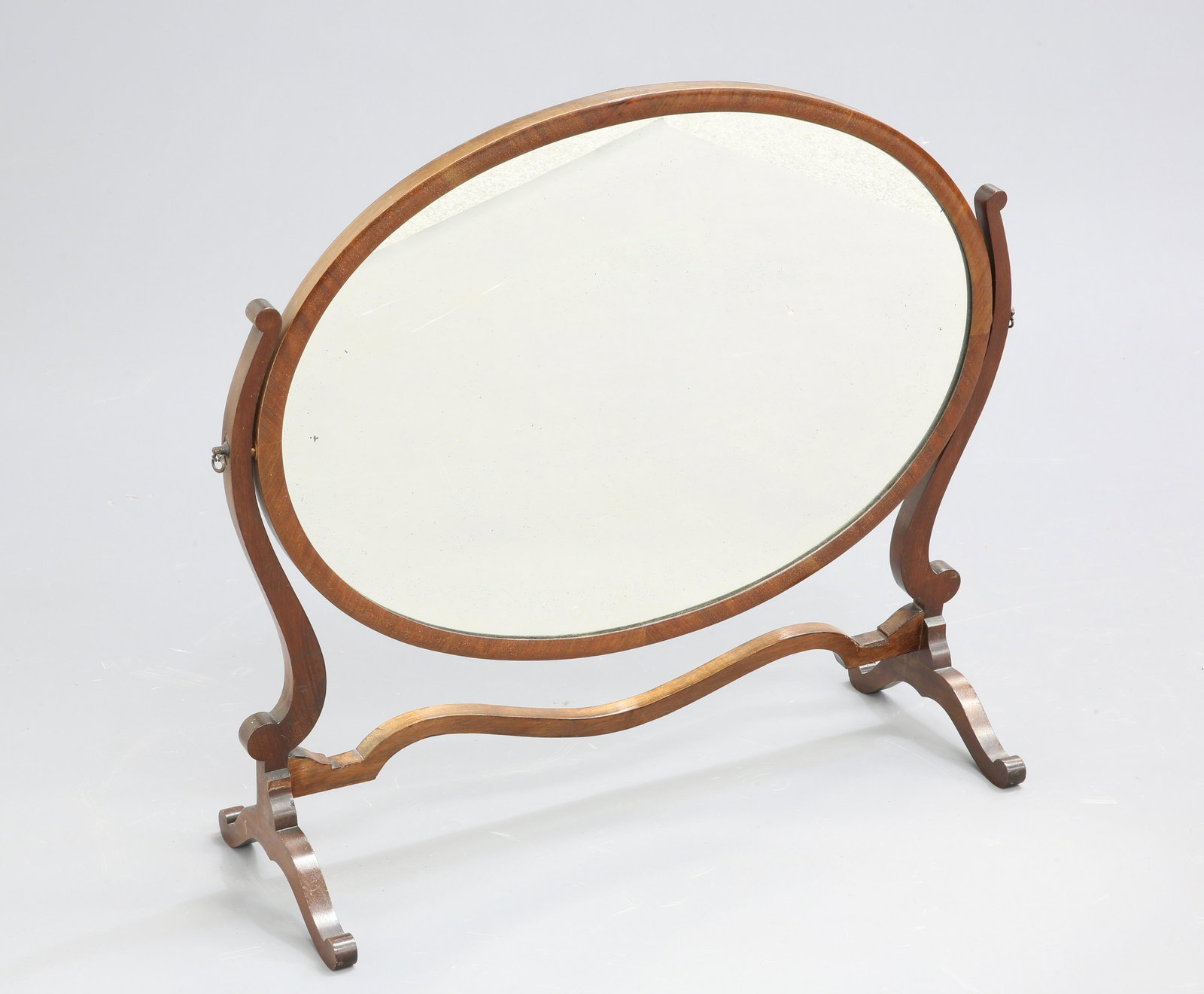 AN EDWARDIAN MAHOGANY OVAL DRESSING TABLE MIRROR, the: AN EDWARDIAN MAHOGANY OVAL DRESSING TABLE MIRROR, the large mirror plate swivelling between S-scroll uprights issuing from a trestle type base. 70cm high, 81cm wide