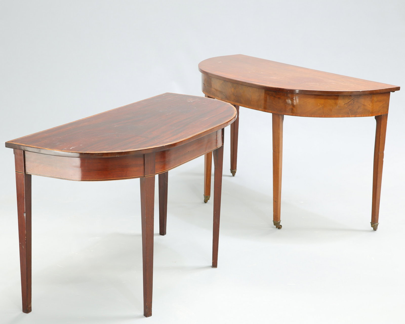 ~ TWO GEORGE III MAHOGANY DEMILUNE SIDE TABLES, the: ~ TWO GEORGE III MAHOGANY DEMILUNE SIDE TABLES, the first with satinwood banded edge and square-sectional tapered legs, the second with satinwood banded frieze and square-sectional tapered legs, movin