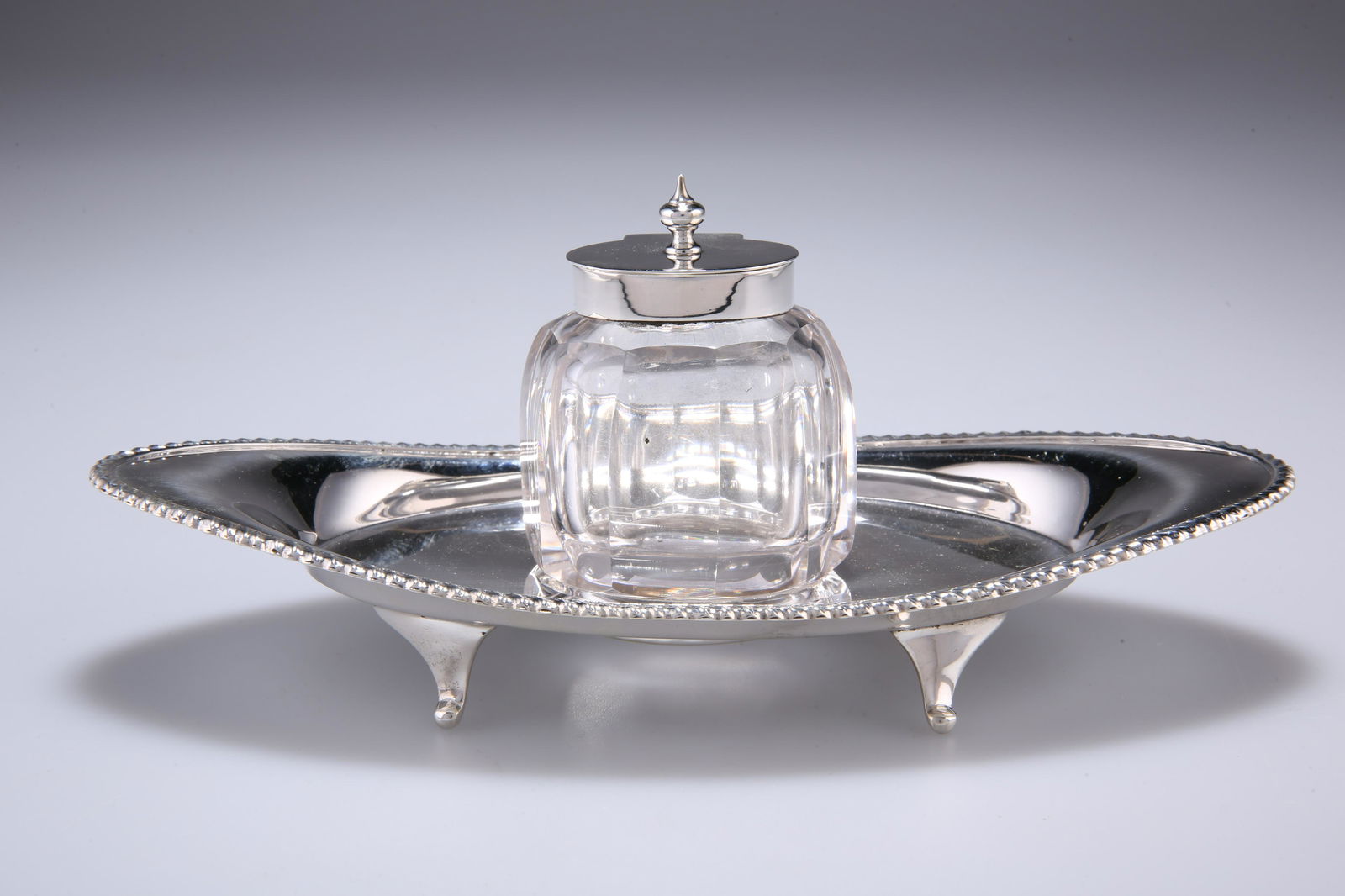 A LATE VICTORIAN SILVER AND CUT GLASS INKWELL AND: A LATE VICTORIAN SILVER AND CUT GLASS INKWELL AND INKSTAND, by William Harrison Walter, Birmingham 1898, the faceted glass inkwell with hinged silver lid topped by a finial, inset to an oval-shaped s