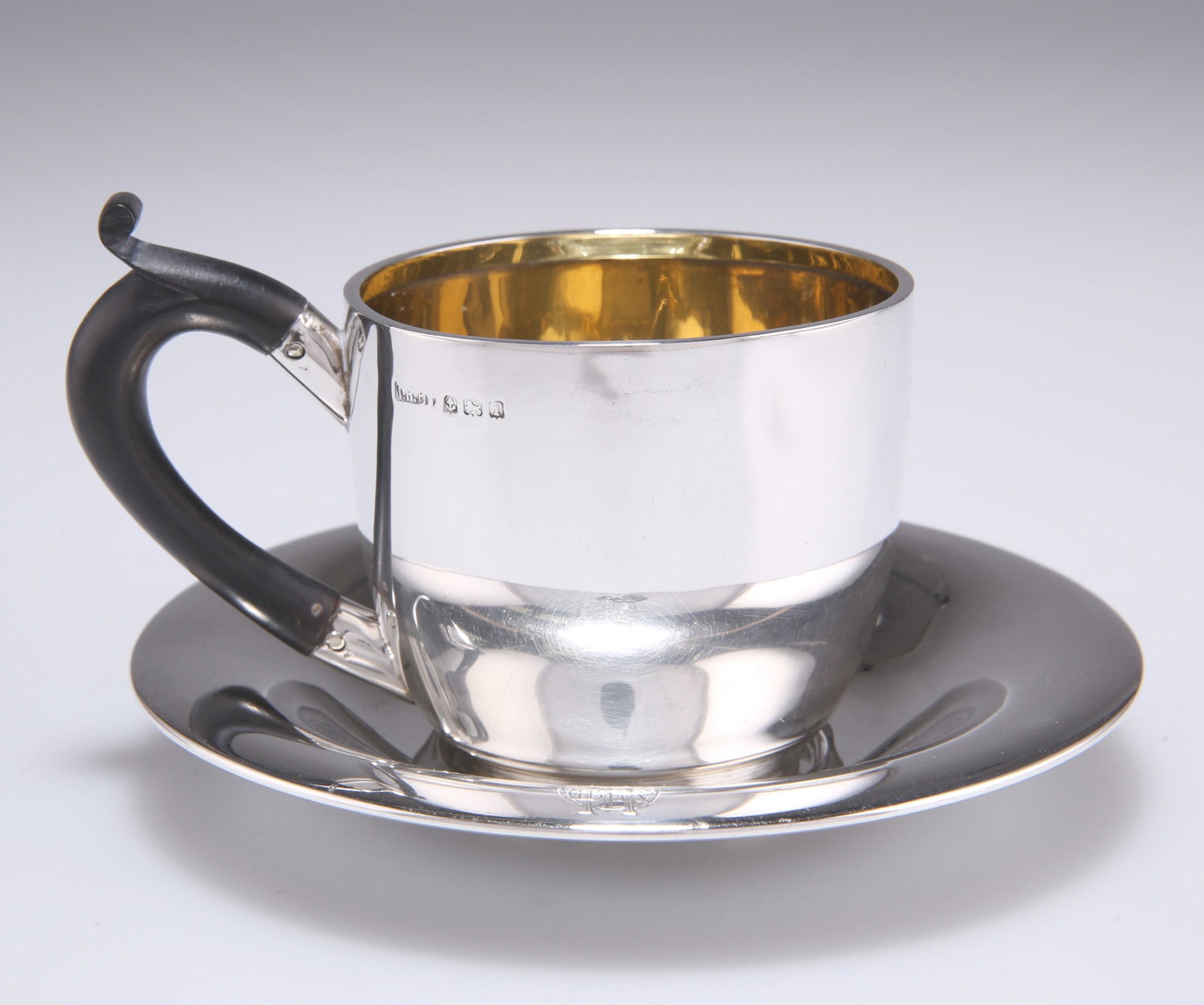 AN EDWARDIAN SILVER CUP AND SAUCER, by T H Hazlewood &: AN EDWARDIAN SILVER CUP AND SAUCER, by T H Hazlewood & Co, Birmingham 1908, plain, the cup with gilded interior and wooden loop handle with scrolling thumbpiece, each engraved with a monogram. (2) S