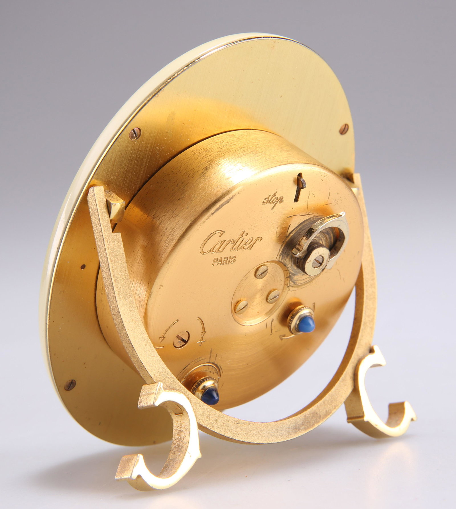 CARTIER: A GILT METAL AND ENAMEL TRAVEL ALARM CLOCK: CARTIER: A GILT METAL AND ENAMEL TRAVEL ALARM CLOCK, oval, with white dial and Roman numerals, within a cream enamel border, on strut stand with twin 'C' feet, two blue stone cabochons to dials on bac