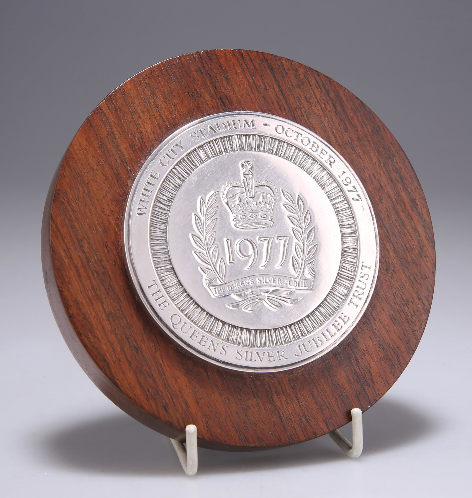GERALD BENNEY: AN ELIZABETH II SILVER-MOUNTED SILVER JUBILEE PAPERWEIGHT: GERALD BENNEY: AN ELIZABETH II SILVER-MOUNTED SILVER JUBILEE PAPERWEIGHT by Gerald Benney, London 1977, the circular hardwood base surmounted by a silver disc inscribed WHITE CITY STADIUM - OCTOBER 19