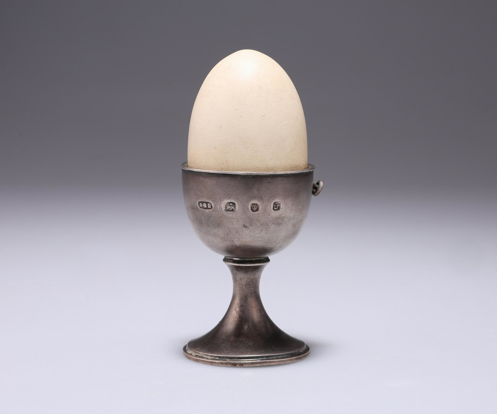 AN ELIZABETH II SILVER NOVELTY EGG TIMER: AN ELIZABETH II SILVER NOVELTY EGG TIMER by Anthony Gordon Elson, London 1980, realistically modelled as a silver egg cup and egg, the removable porcelain-covered egg top reveals a carousel with rabbi
