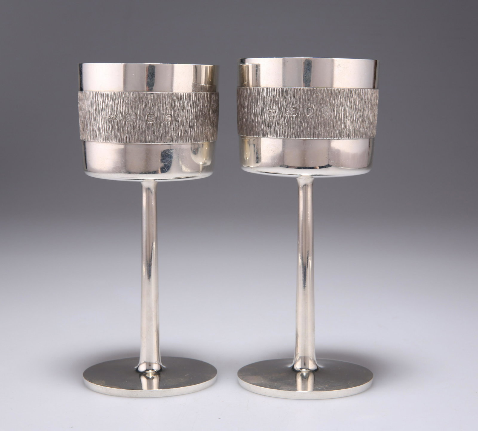 A PAIR OF ELIZABETH II SILVER GOBLETS: A PAIR OF ELIZABETH II SILVER GOBLETS maker R A F, London 1971, the straight sided bowls with a central girdle of bark effect, gilded to interior, each raised on a straight stem issuing from a flat ci
