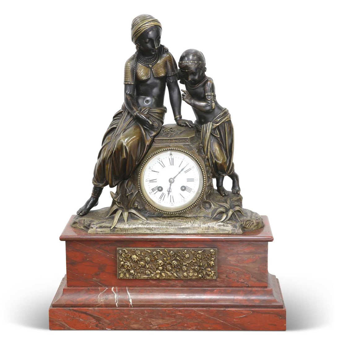 A 19TH CENTURY BRONZE AND RED MARBLE GARNITURE DE CHEMINEE: A 19TH CENTURY BRONZE AND RED MARBLE GARNITURE DE CHEMINEE the clock modelled as a mother and child standing on a naturalistic ground encasing a two-train drum clock movement, the dial with Roman num