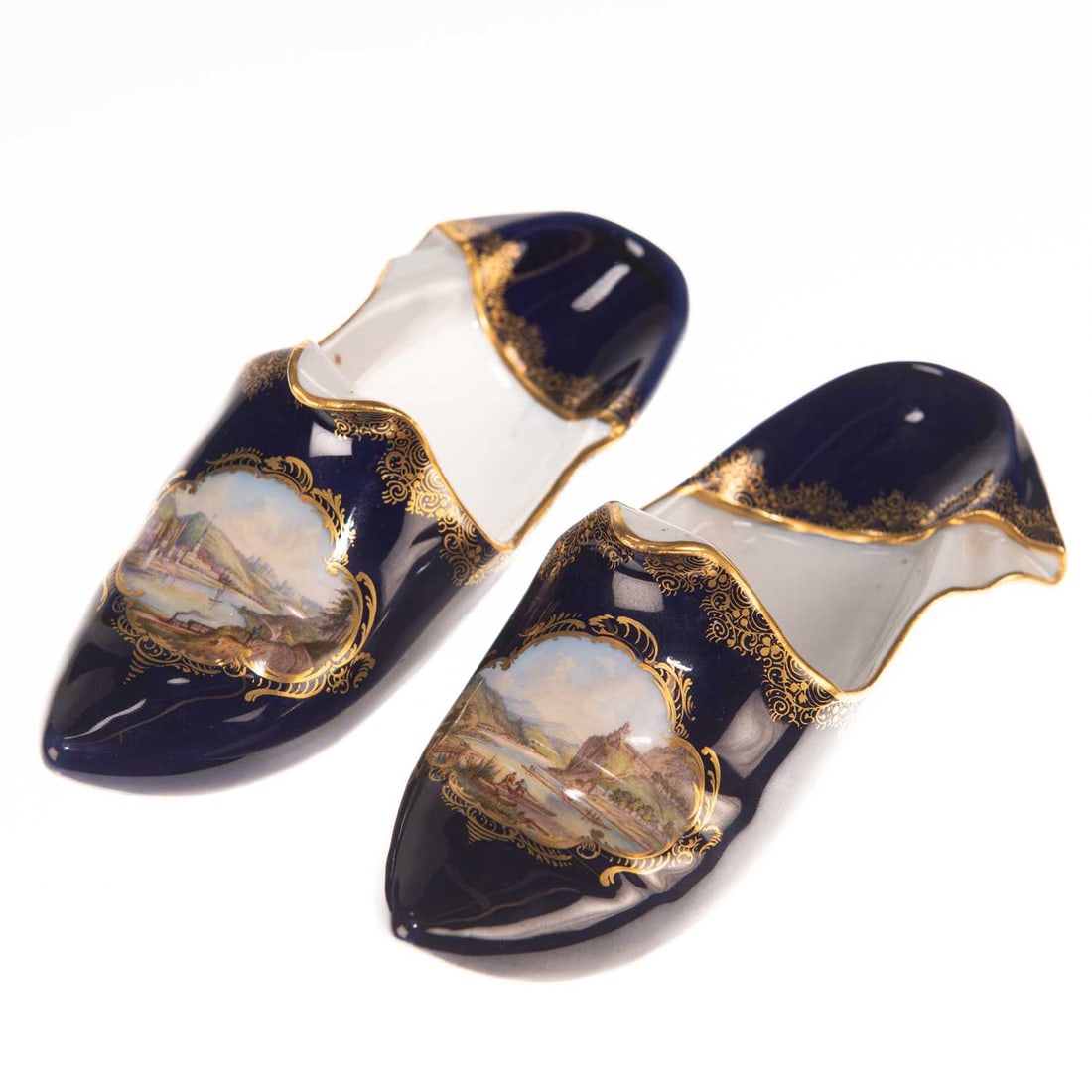A PAIR OF MEISSEN BLUE-GROUND MODELS OF SLIPPERS: A PAIR OF MEISSEN BLUE-GROUND MODELS OF SLIPPERS each painted with a river landscape within a quatrefoil panel, highlighted with gilding, titled on the undersides, underglaze blue crossed swords. (2) 