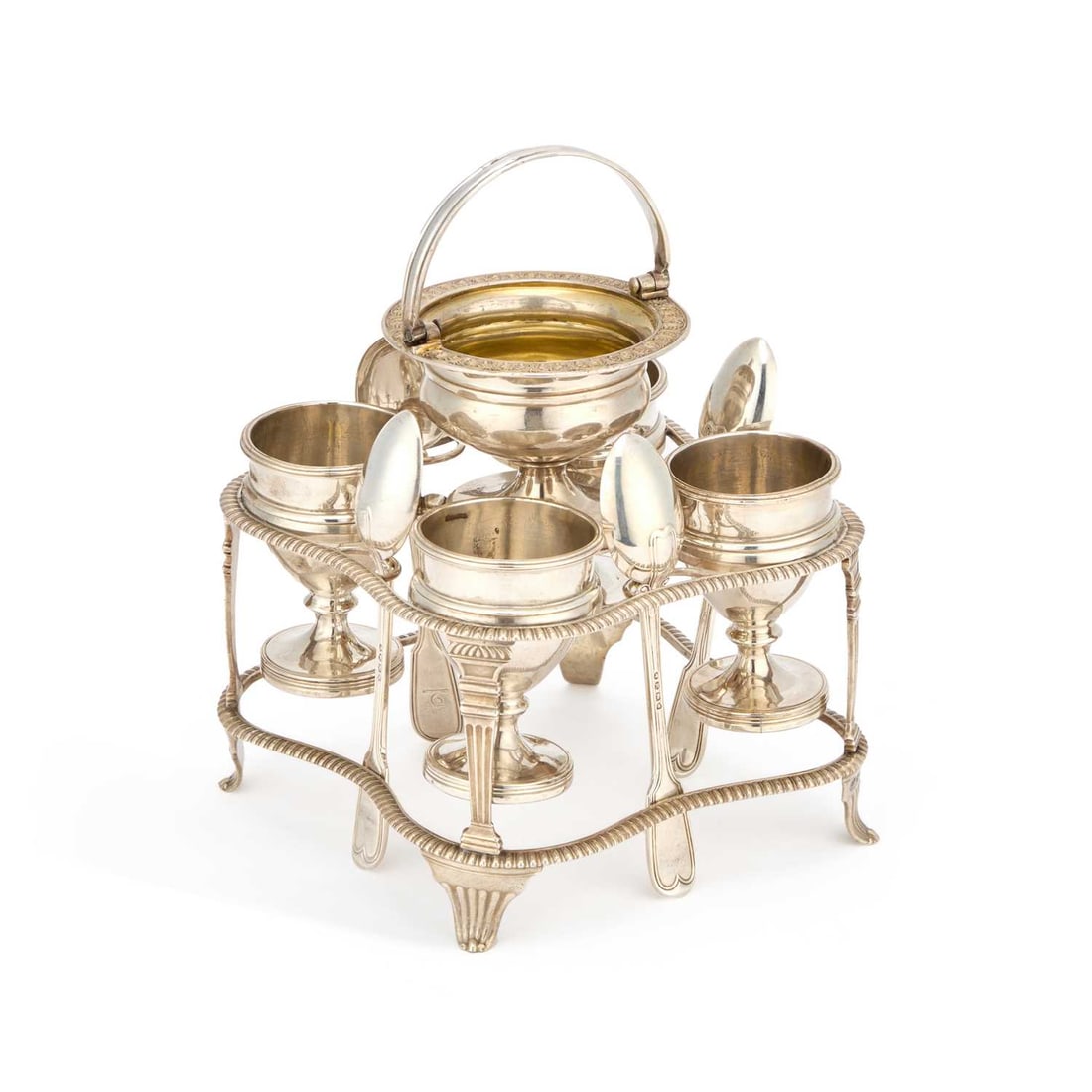 A GEORGE III SILVER EGG CRUET: A GEORGE III SILVER EGG CRUET by John Emes, London 1807, of shaped square form with gadrooned rims, the four egg cups with applied girdles seated in circular reserves, surmounted by a swing-handle urn