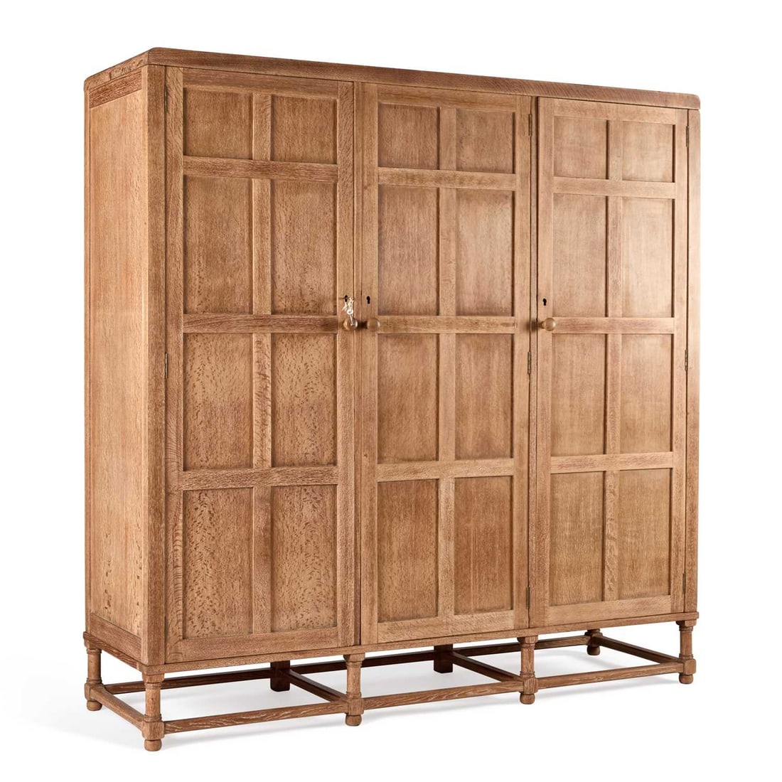 AMBROSE HEAL FOR HEAL'S, A LIMED OAK TRIPLE WARDROBE EARLY 20TH CENTURY: AMBROSE HEAL FOR HEAL'S, A LIMED OAK TRIPLE WARDROBE EARLY 20TH CENTURY with three eight-panel doors, raised on a block and columnar stand, the end sections with rails and hooks for hanging, the centr